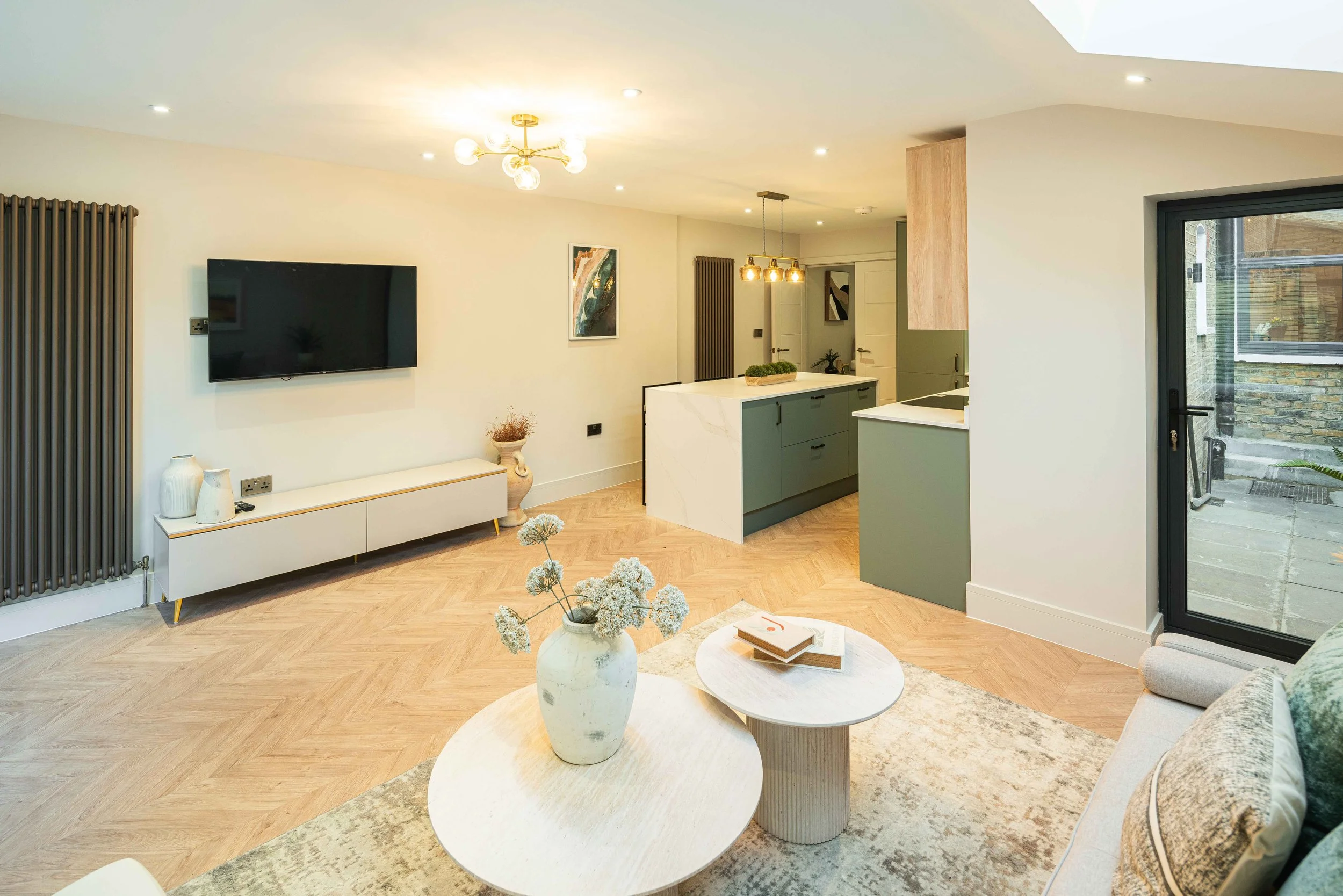 Modern open-plan living room with a wall-mounted TV, a white media unit, and minimalist decor, connected to a kitchen with green cabinetry, a marble island, and pendant lighting, with a sliding glass door leading outdoors.