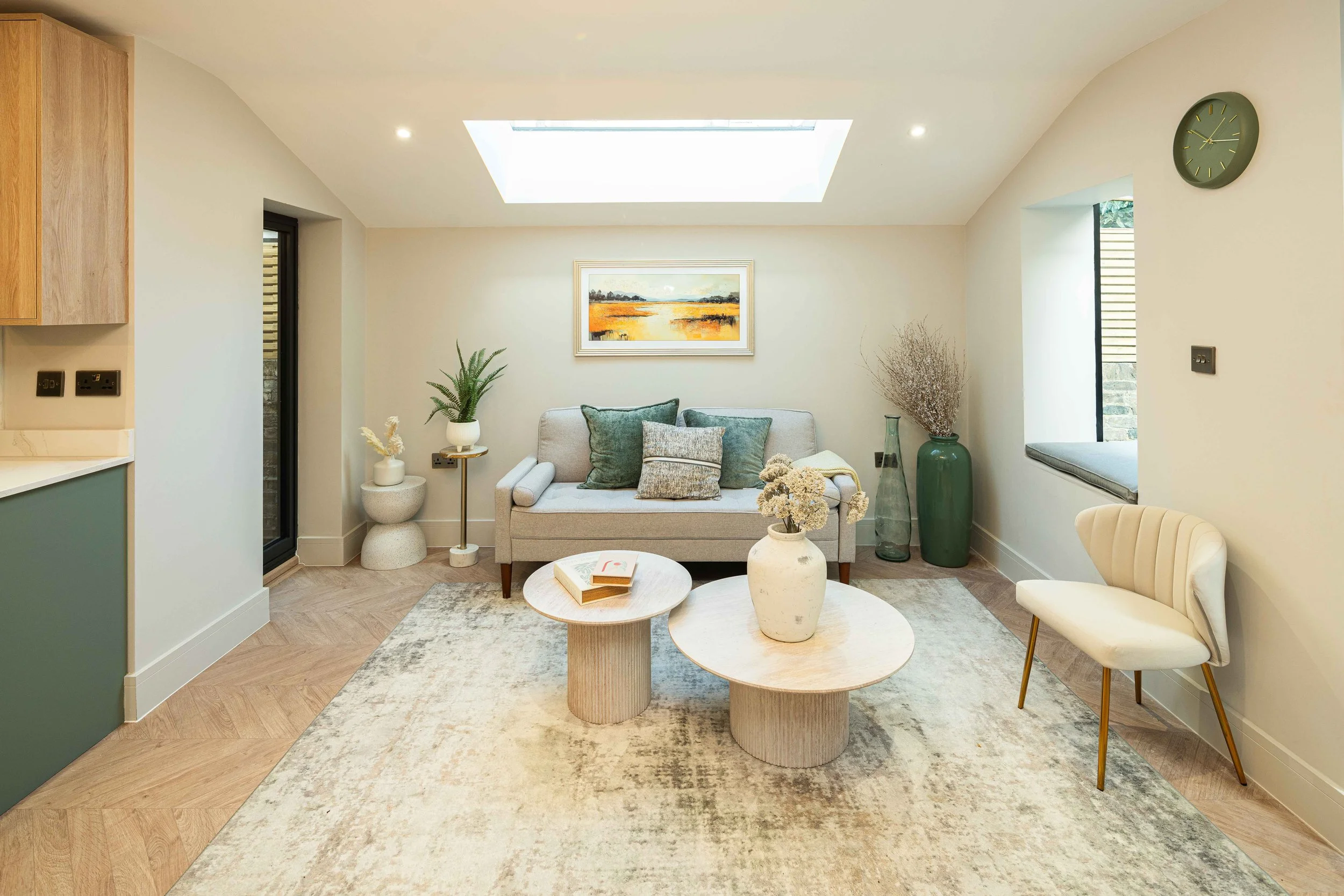 Living room with a beige sofa, decorative cushions, a coffee table with a vase of flowers, two green vases, a wall clock, a window seat, and a framed landscape painting, illuminated by natural light from a large skylight.