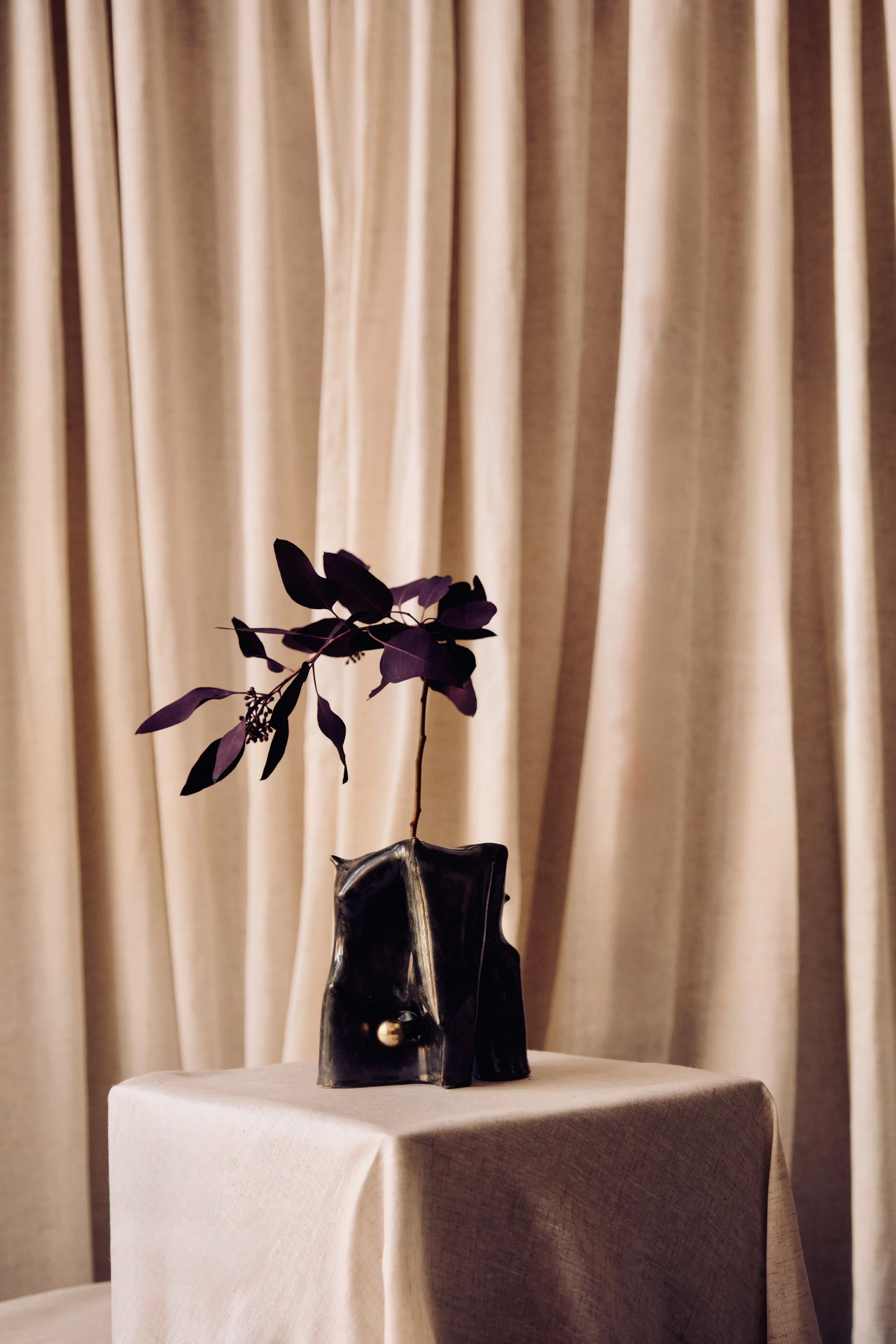 Schwarze Vase mit lila Blättern und kleinen Blumen auf einem Tisch vor einem beige Vorhang.