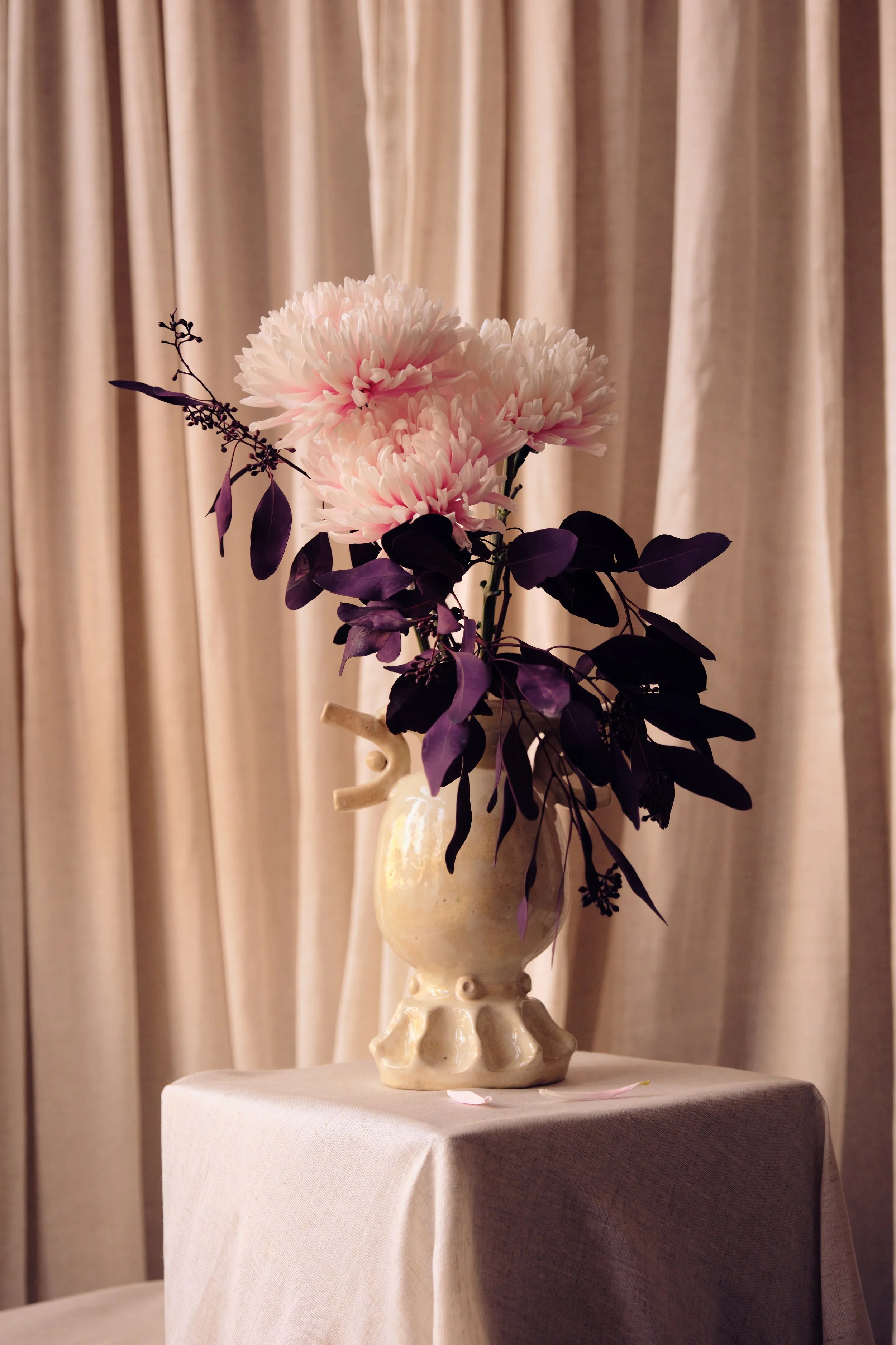 Blumenstrauß mit weißen und rosa Blüten in einer cremefarbenen Vase auf einem Tisch vor cremefarbenen Vorhängen.