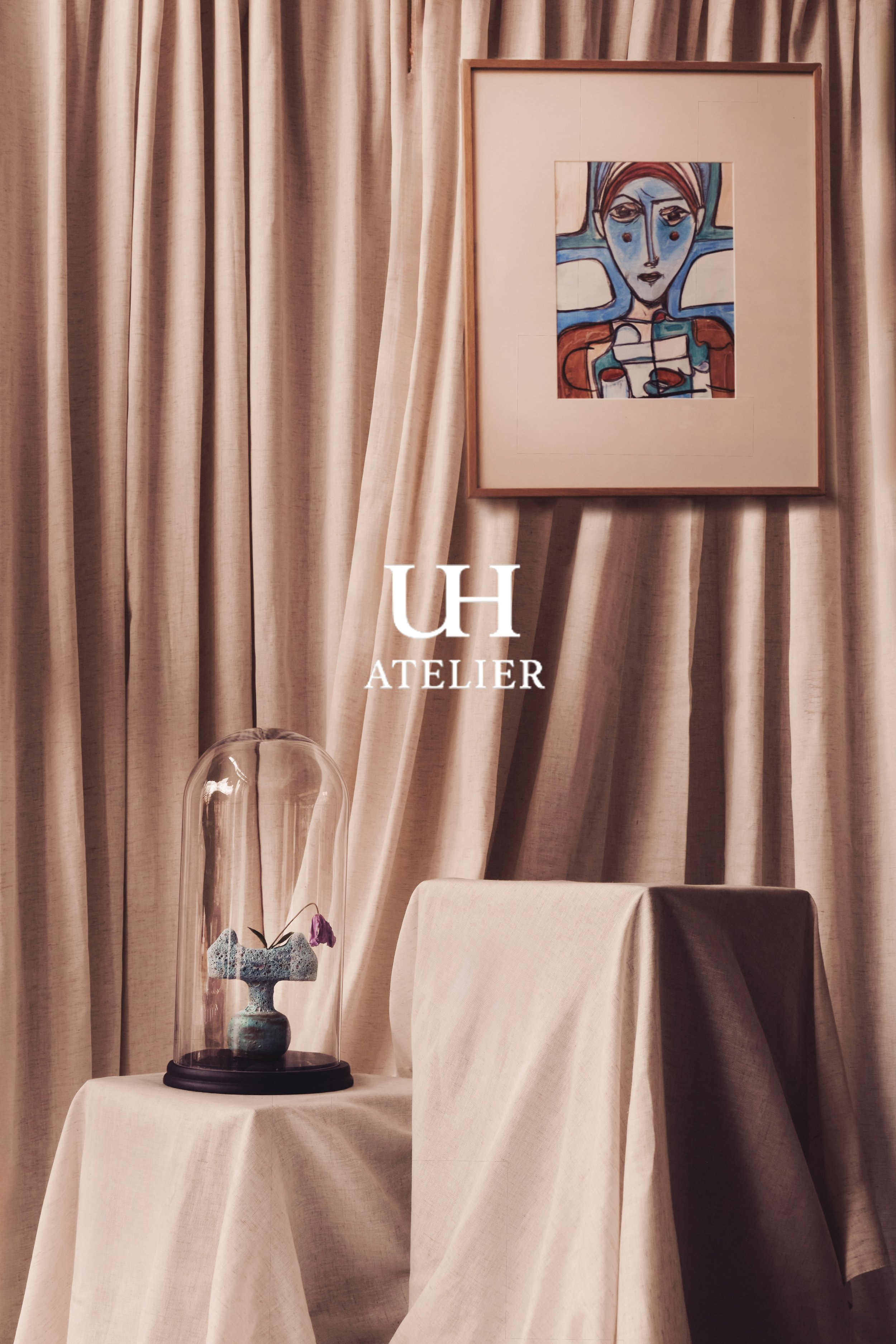Ein Bild mit einem beige Vorhang, einem gerahmten abstrakten Gemälde an der Wand, einem Kleinformat auf einem Tisch und einer festen Glaskuppel über einer Kunstinstallation. Der Schriftzug 'UH ATELIER' ist auf dem Bild sichtbar.