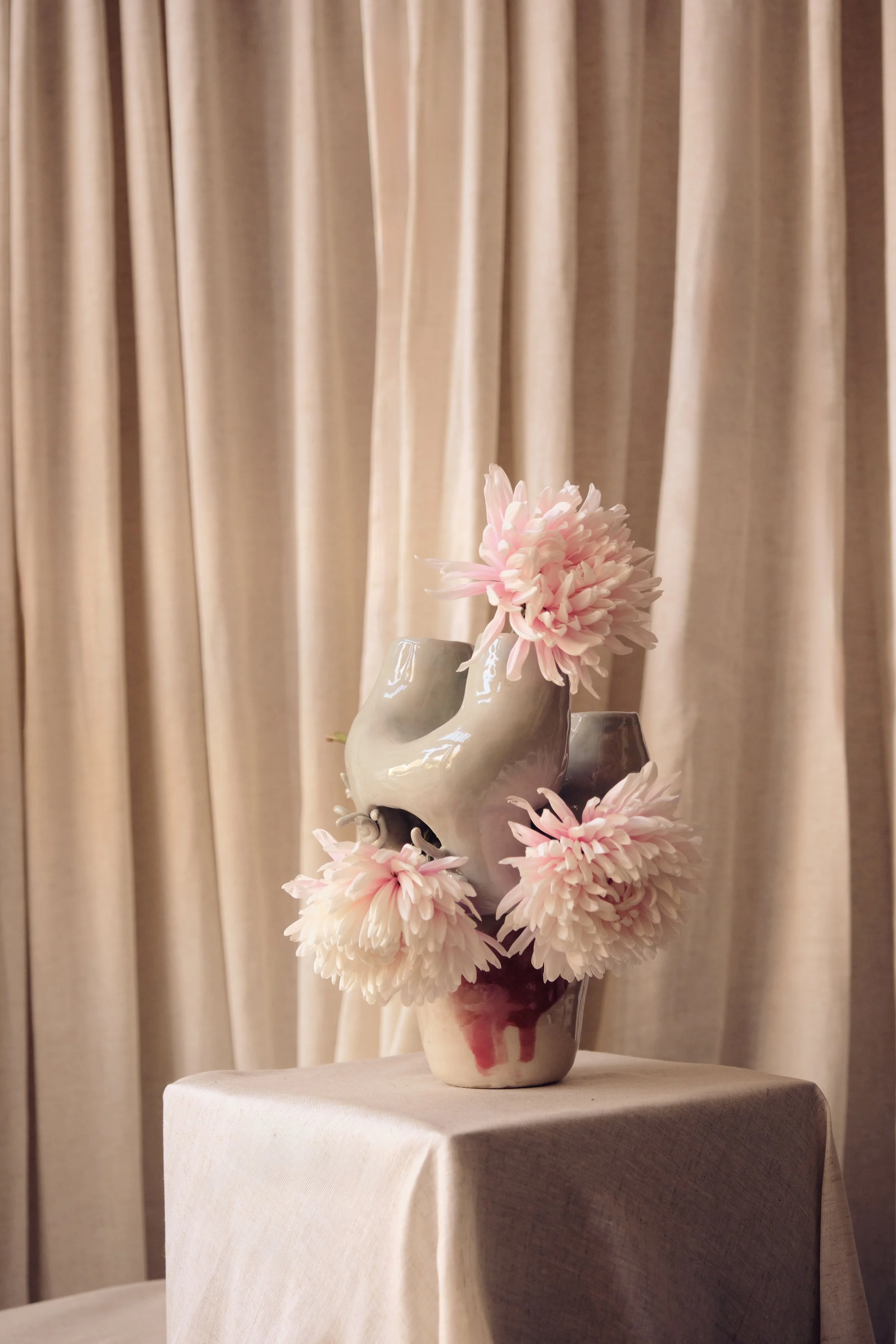 Eine weiße Keramikvase mit rosa Blumen auf einem Tisch vor einem beige Vorhang.