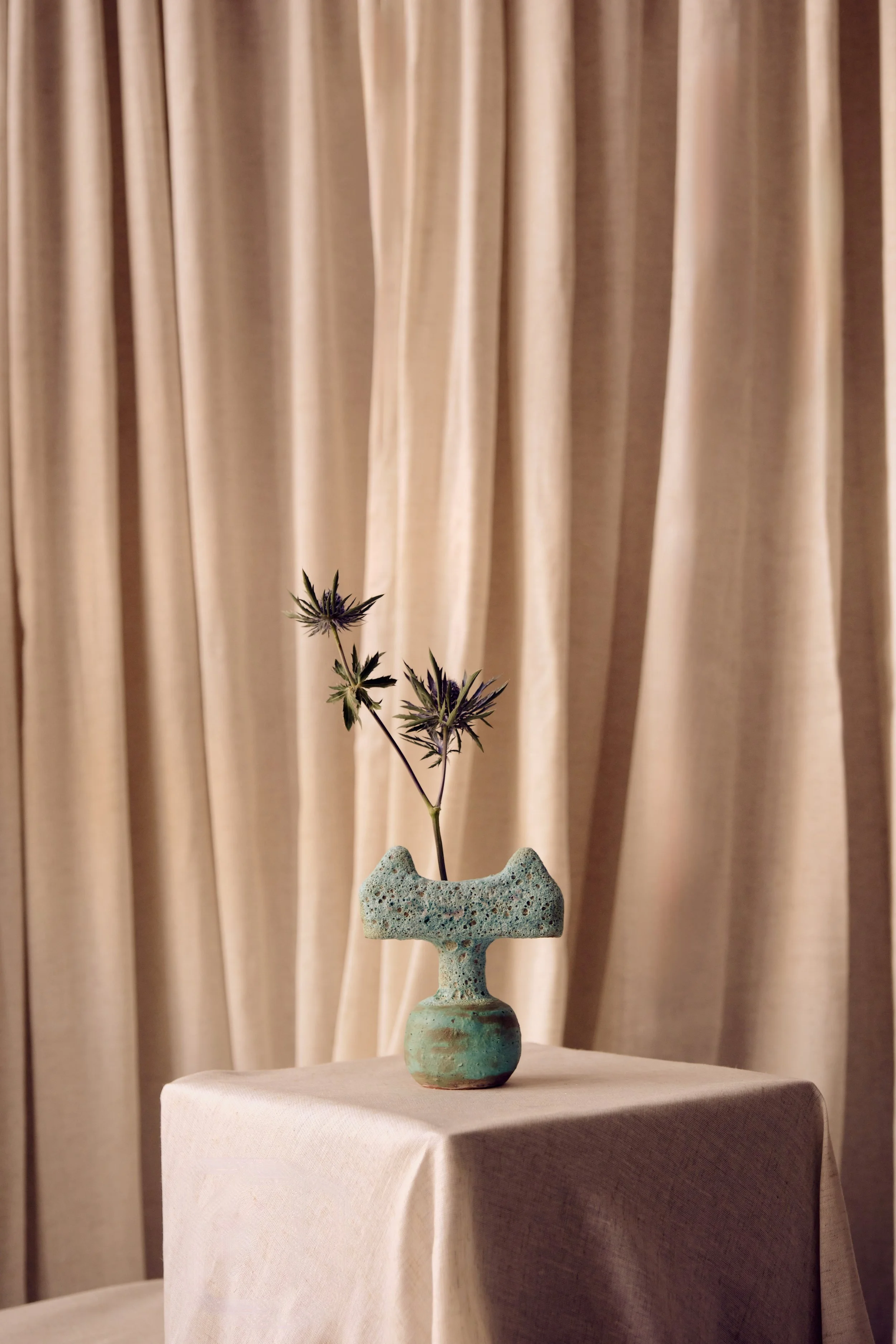 Eine grüne Keramikvase mit Blume, vor einem beige Vorhang.