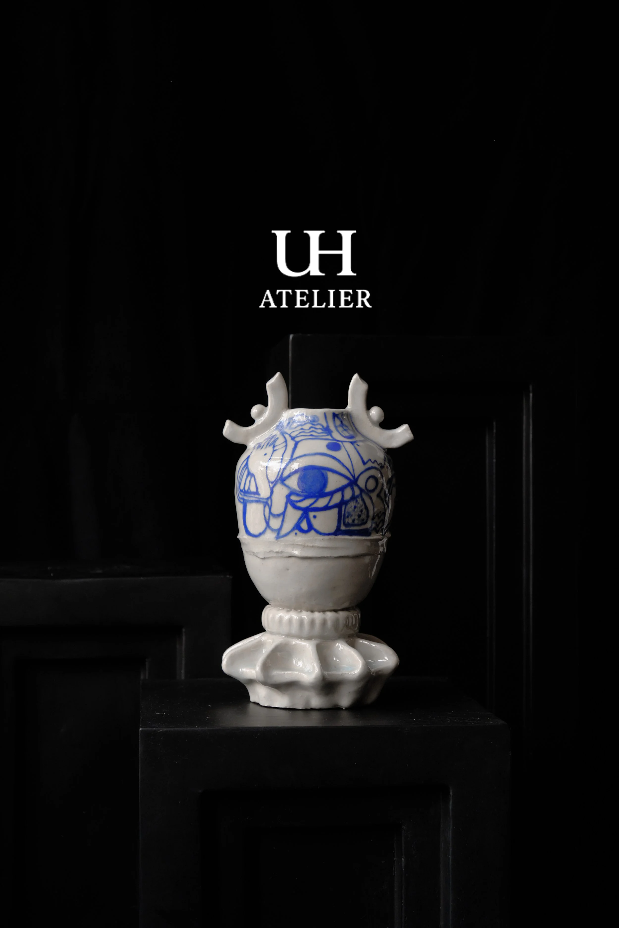 Ein weißer Keramikvase mit blauer Verzierung, auf einem schwarzen Sockel, im Hintergrund ein schwarzer Raum mit der Beschriftung 'UH ATELIER' in Weiß.