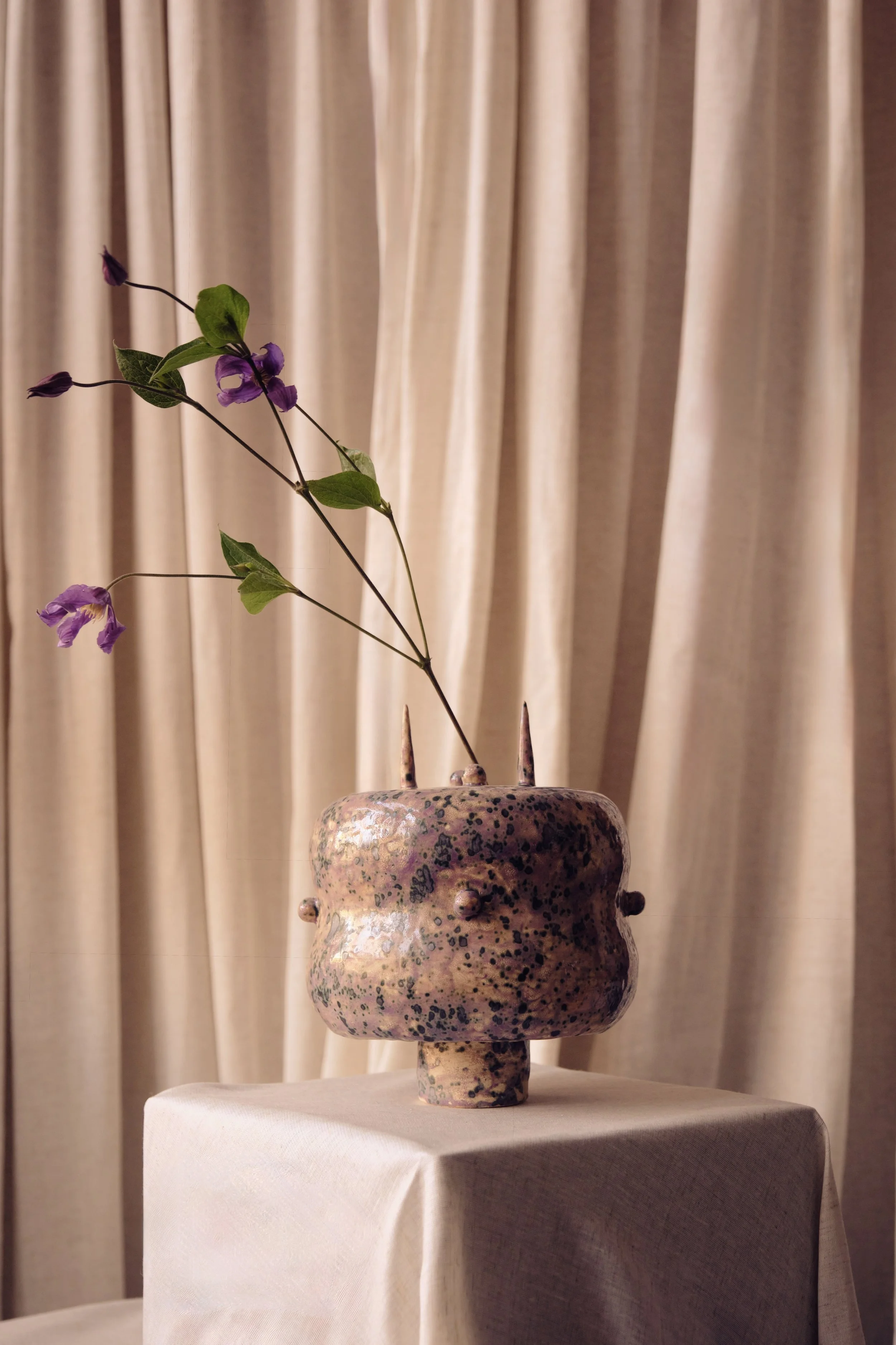 Keramikvase mit Blumen auf einem weißen Tisch vor einer beige Gardine.