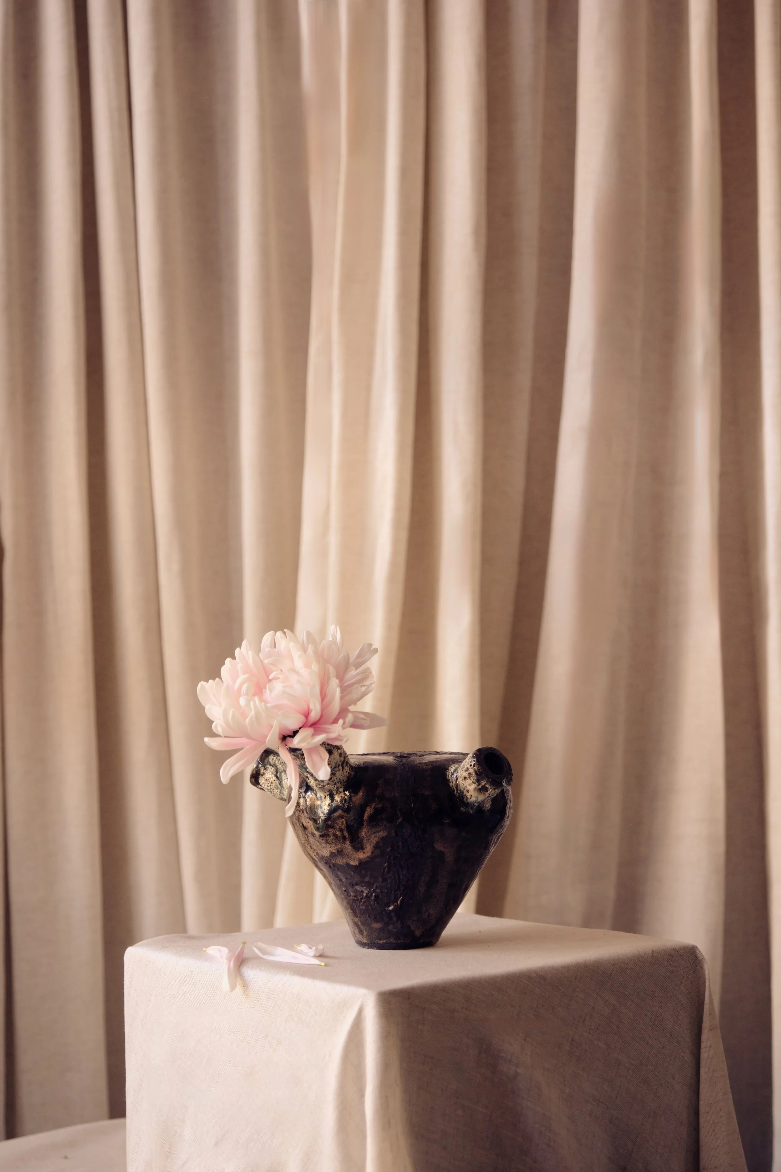 Eine Keramikvase mit einer pinken Blumen auf einem Tisch vor beige Vorhängen.