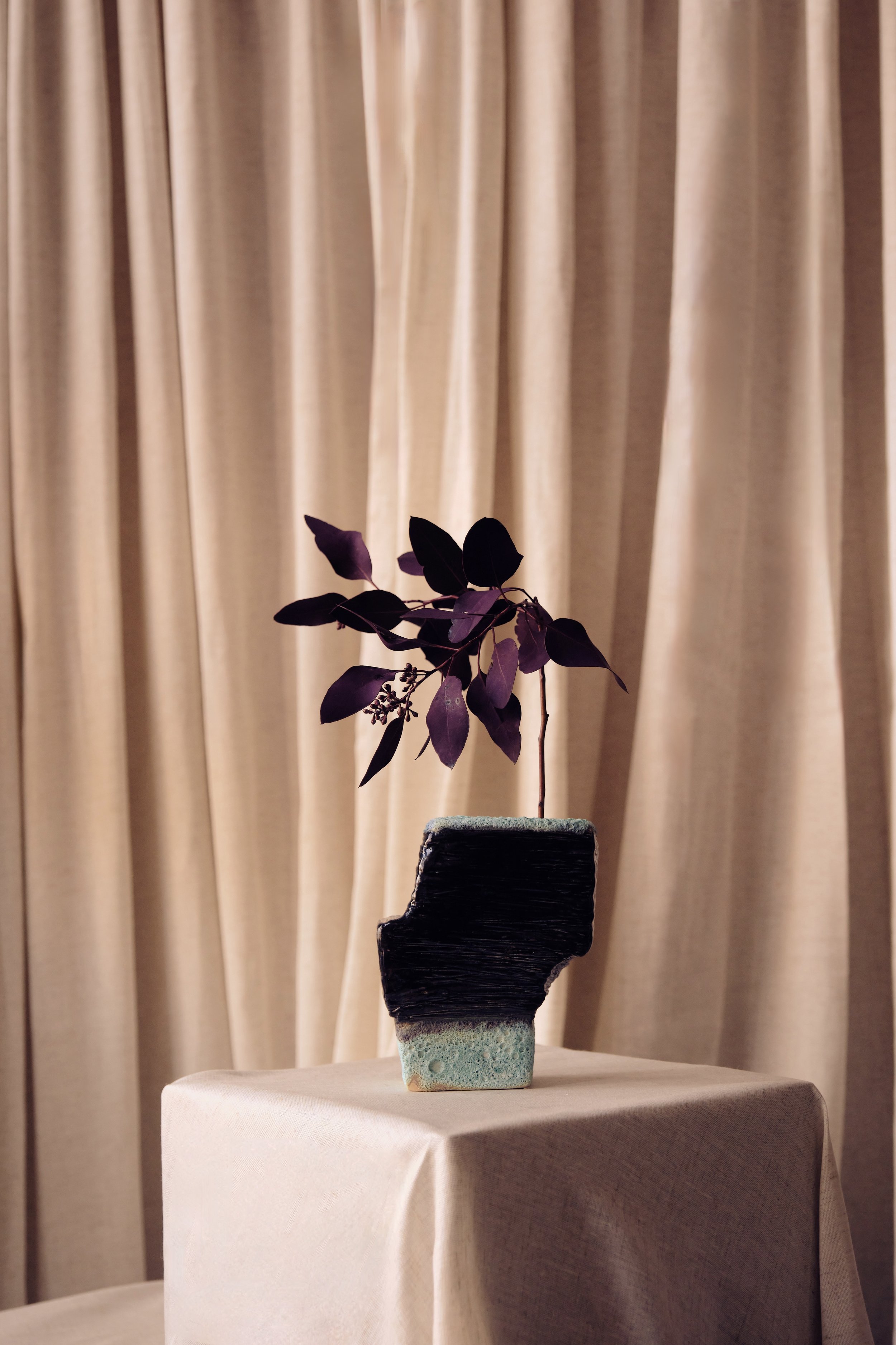 Eine Vase mit lilafarbenen Blättern auf einem weißen Tisch vor beigen Gardinen.