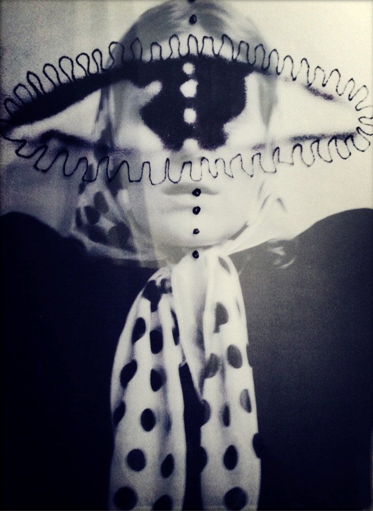 Schwarz-weiß Bild einer Person mit einem großen, geflochtenen Hut mit Mustern und einer polka-dot-schleife. Der Hut hat eine Zierde auf der Stirn und das Oberteil der Person ist mit einem gepunkteten Tuch bedeckt.