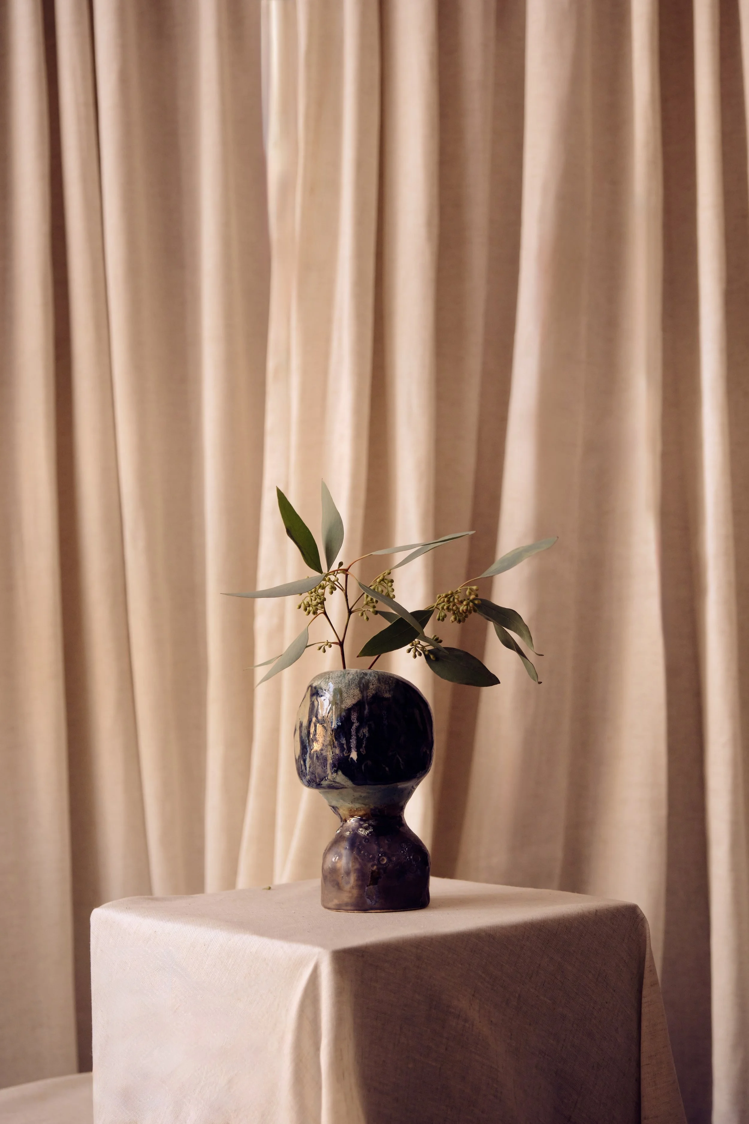 Blumen in einer Keramikvase auf einem weißen Tisch vor einem Beigefarbenen Vorhang.