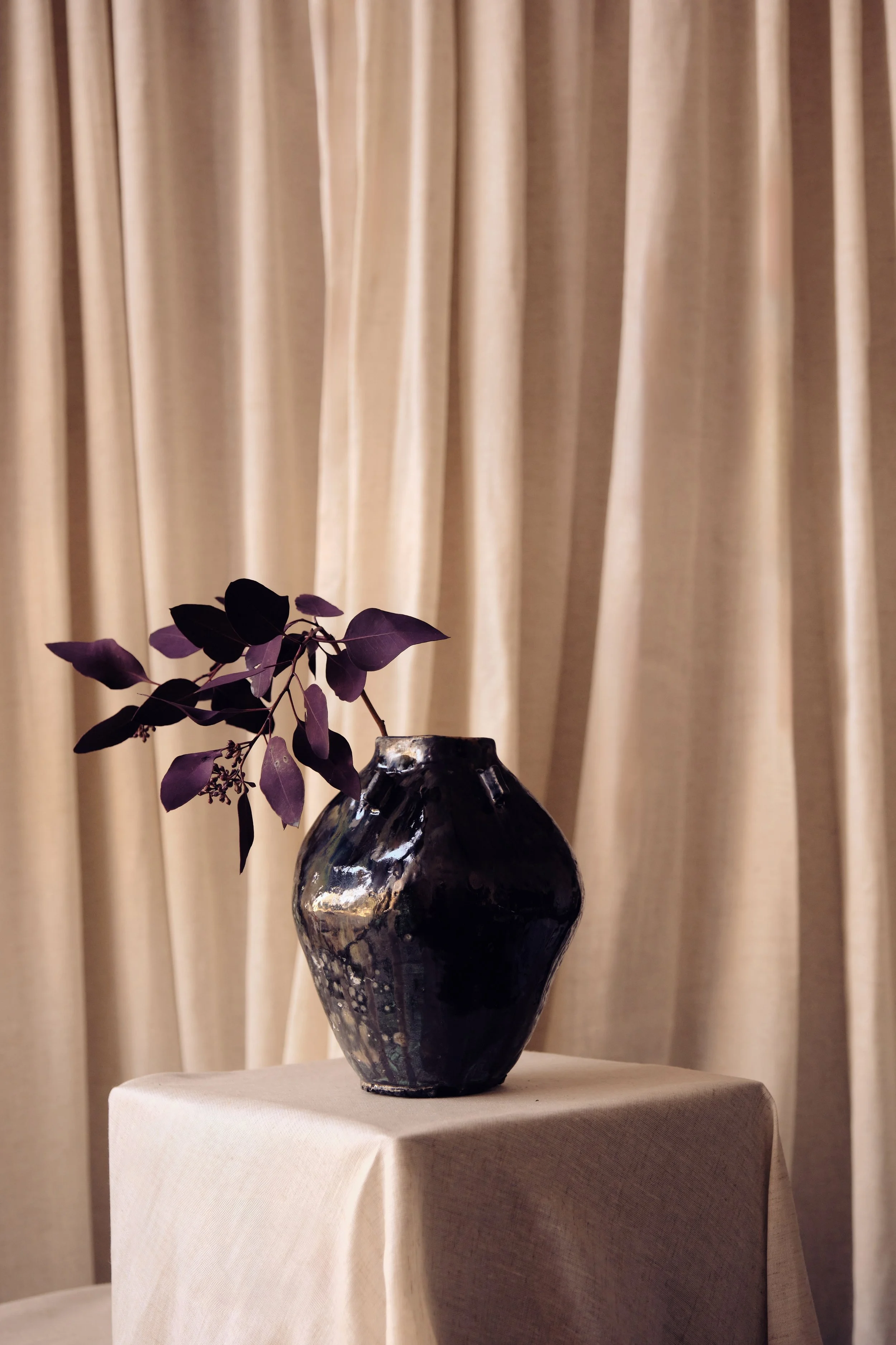 Eine dunkle, metallisch reflektierende Vase mit violetten Blättern und kleinen Beeren auf einem weißen Tischtuch, vor einem beige Stoffvorhang.