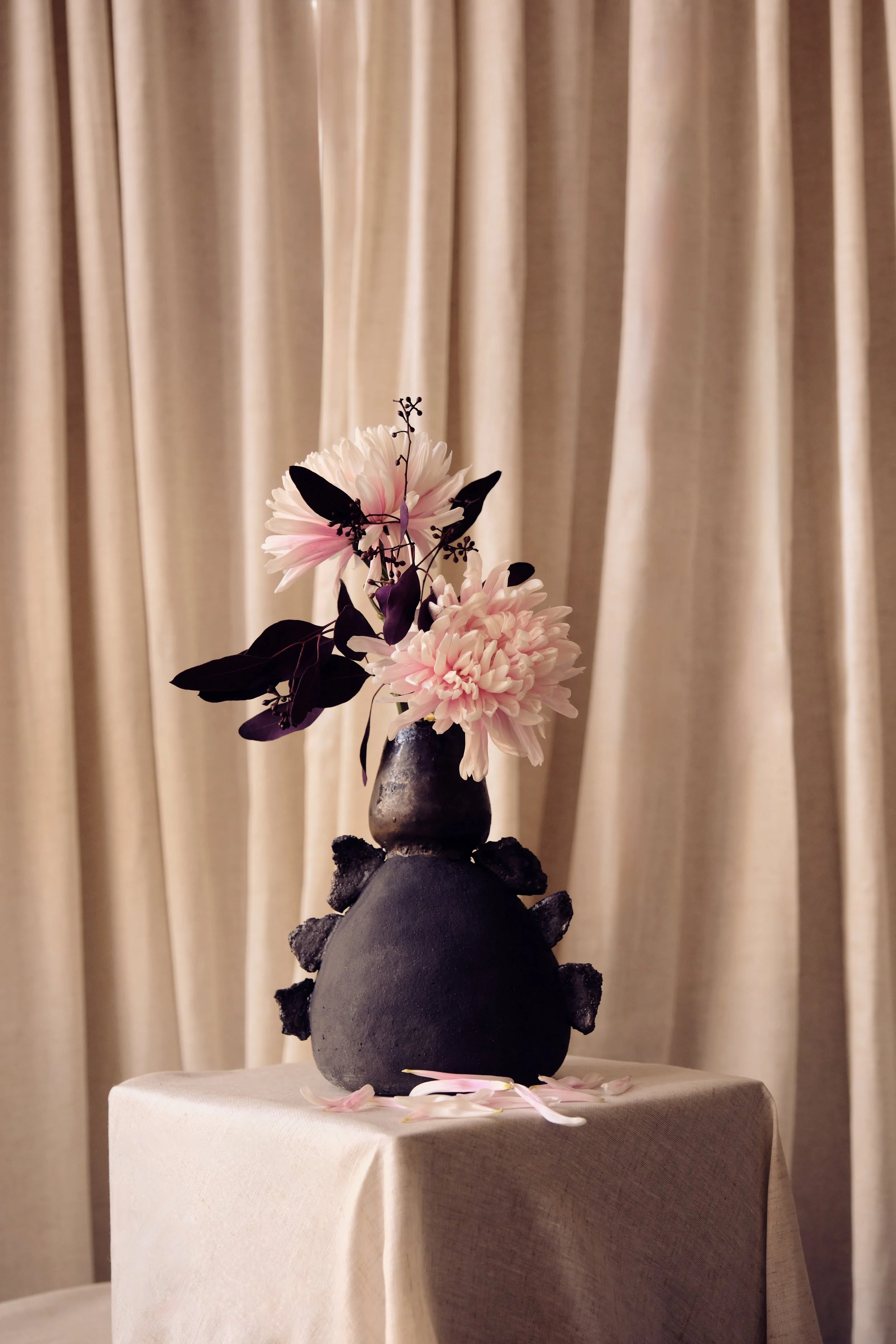 Blumenvase mit pinken und violetten Blumen auf einem Tisch vor beigen Vorhängen.
