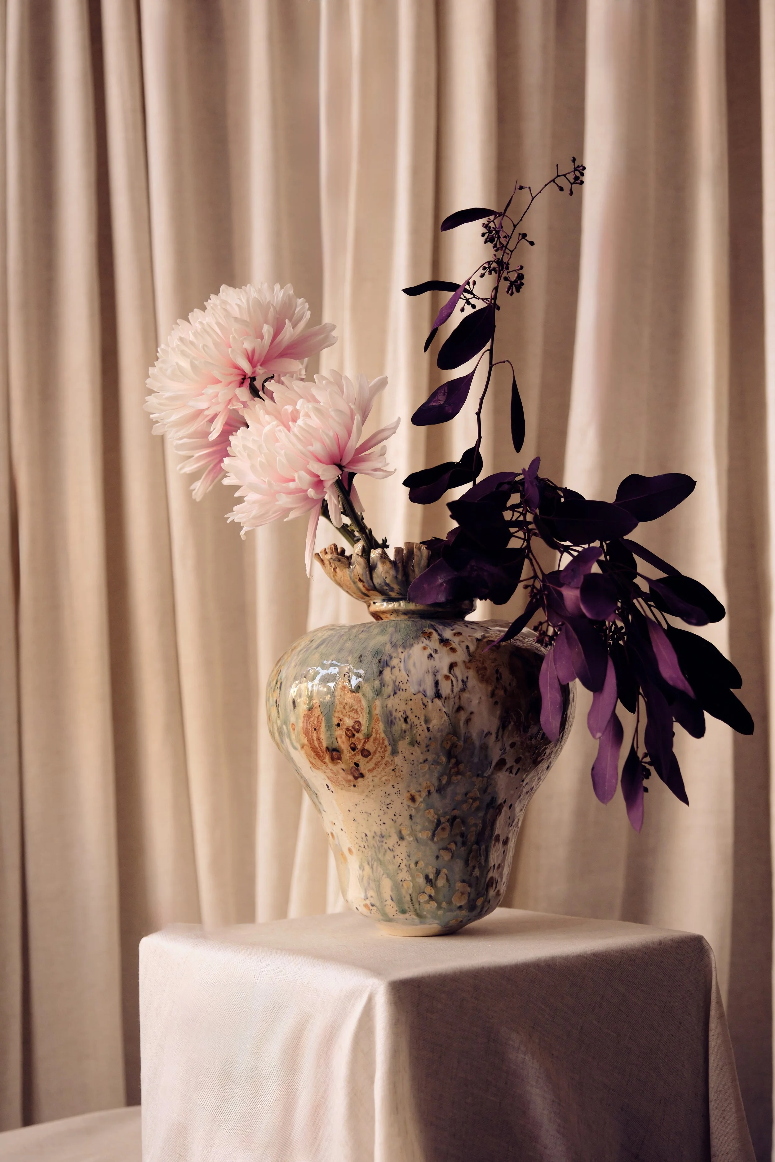 Eine bunte Keramikvase mit weißen und rosa Blumen und violetten Blättern, auf einem weißen Tuch auf einem Tisch mit einem beige-farbigen Vorhang im Hintergrund.