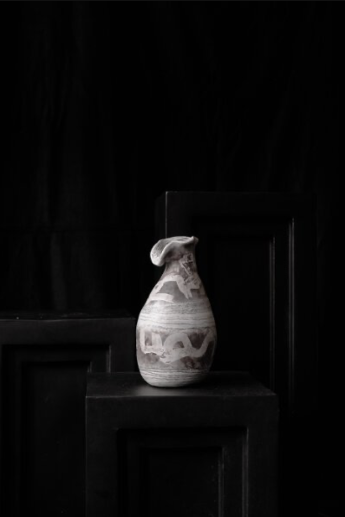 Eine tassenartige Keramikvase mit einer weißen und braunen Marmorierung steht auf einem schwarzen Podest vor einem schwarzen Hintergrund.