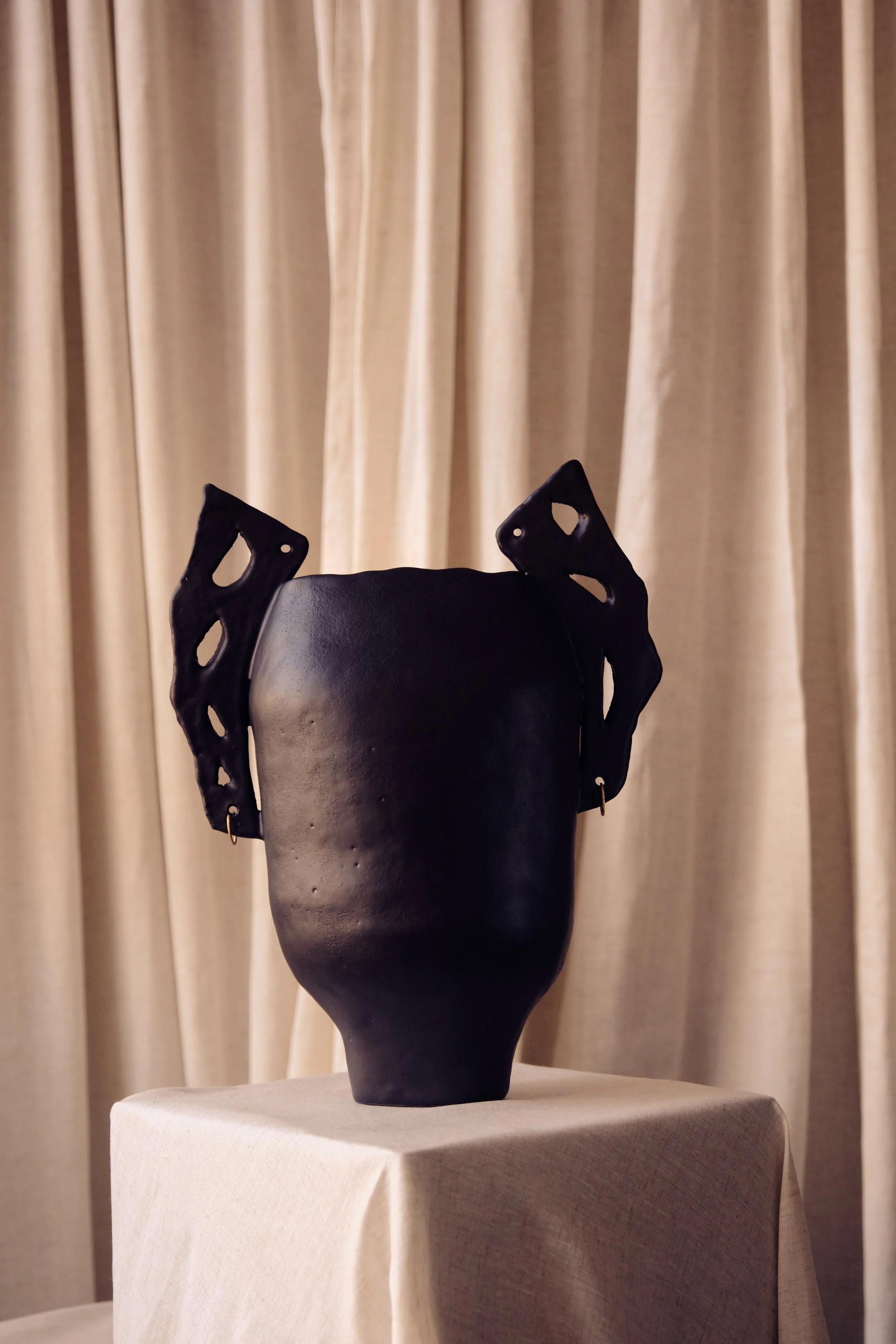 Schwarze Skulptur eines menschlichen Gesichts auf einem Standfuß vor einem beige-farbenen Vorhang.