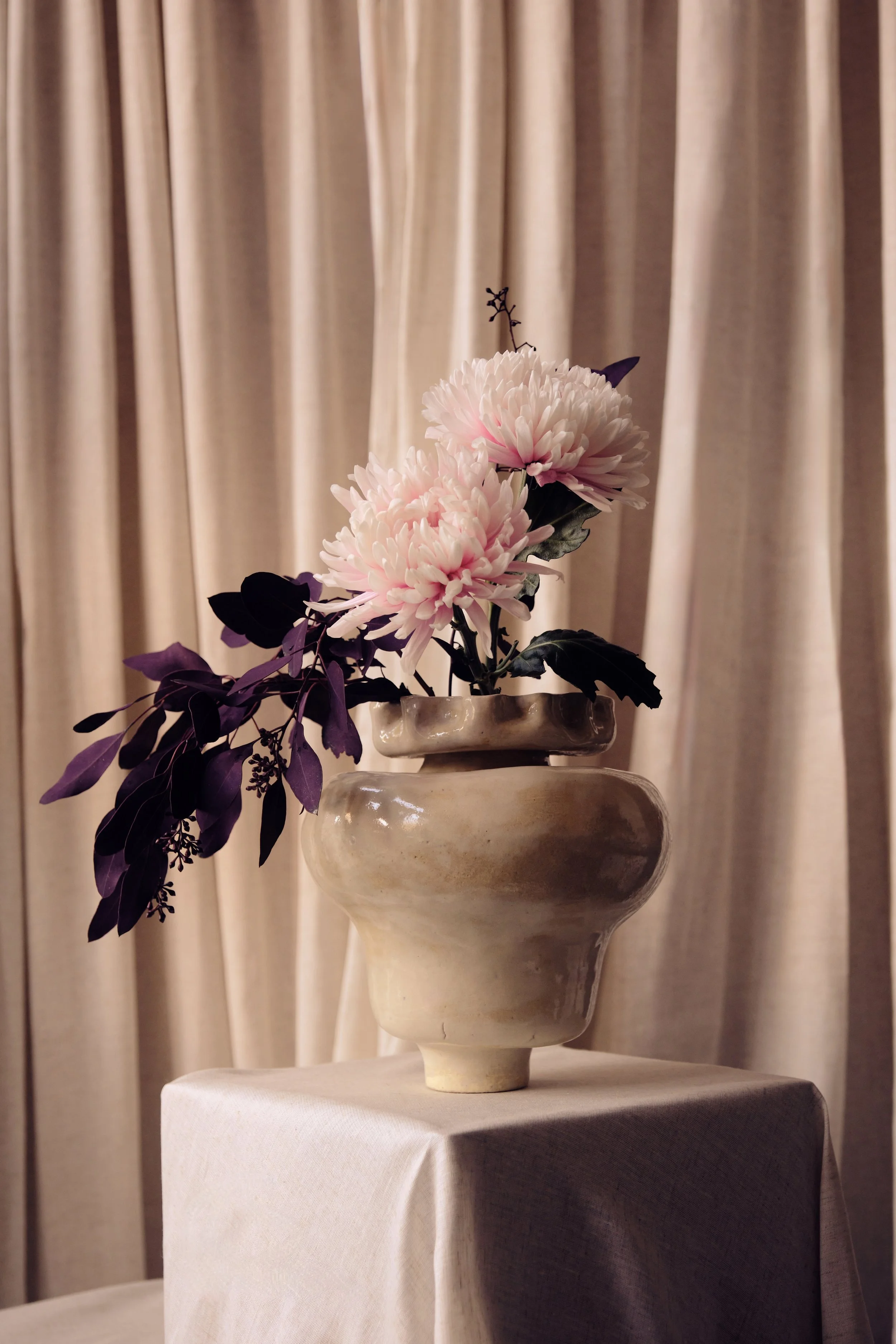 Eine Vase mit zwei großen, weißen und rosafarbenen Blumen und dunklen Blättern steht auf einem weißen Tisch vor einem beige-rosafarbenen Vorhang.