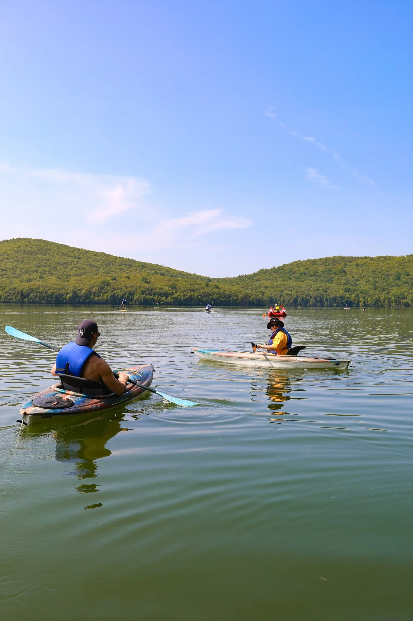People kayaking on a calm lake with green hills and a blue sky in the background.
