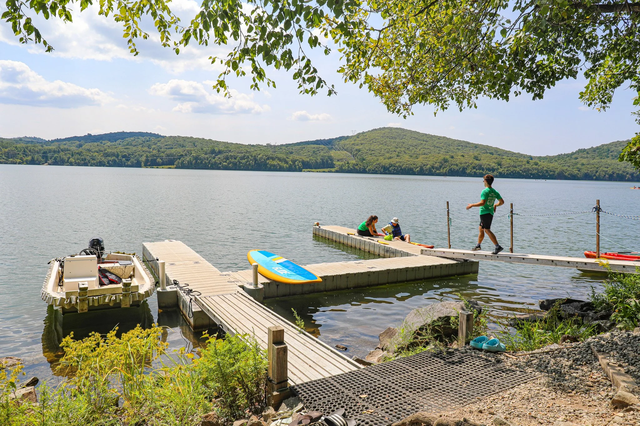 People at a lakeside dock with boats and a paddleboard, surrounded by green hills and a cloudy sky.