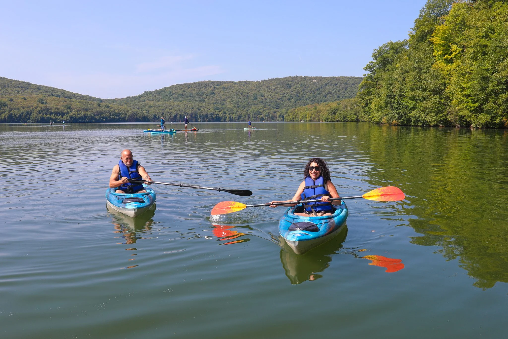Two people kayaking on a lake with forested hills in the background, and several paddleboarders in the distance.