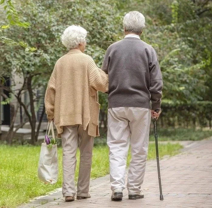 An elderly couple walking together in a park, man using a cane for support, woman carrying a tote bag.