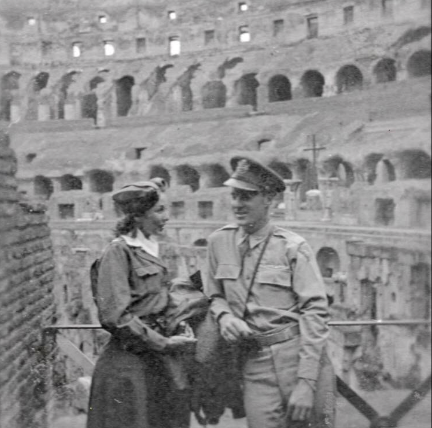 A black and white photograph of a man in military uniform and a woman in vintage clothing, standing together at the Colosseum in Rome.