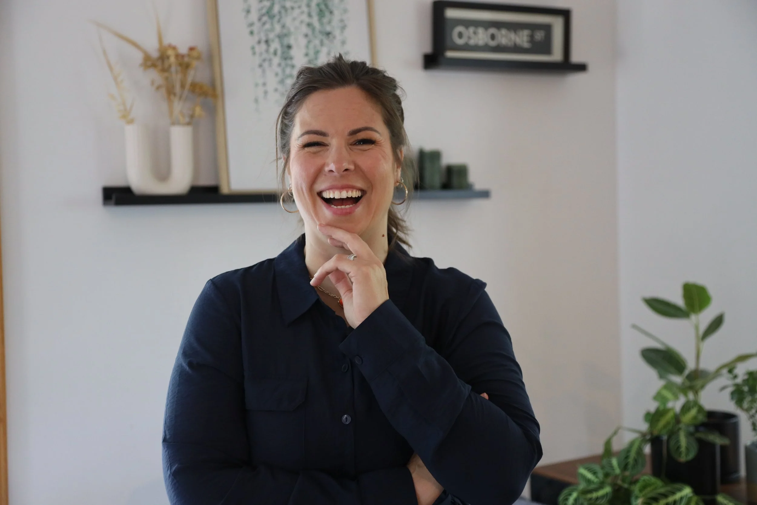 A woman laughing with her hand on her chin, wearing a dark blue shirt, in a modern indoor setting with houseplants and wall art.