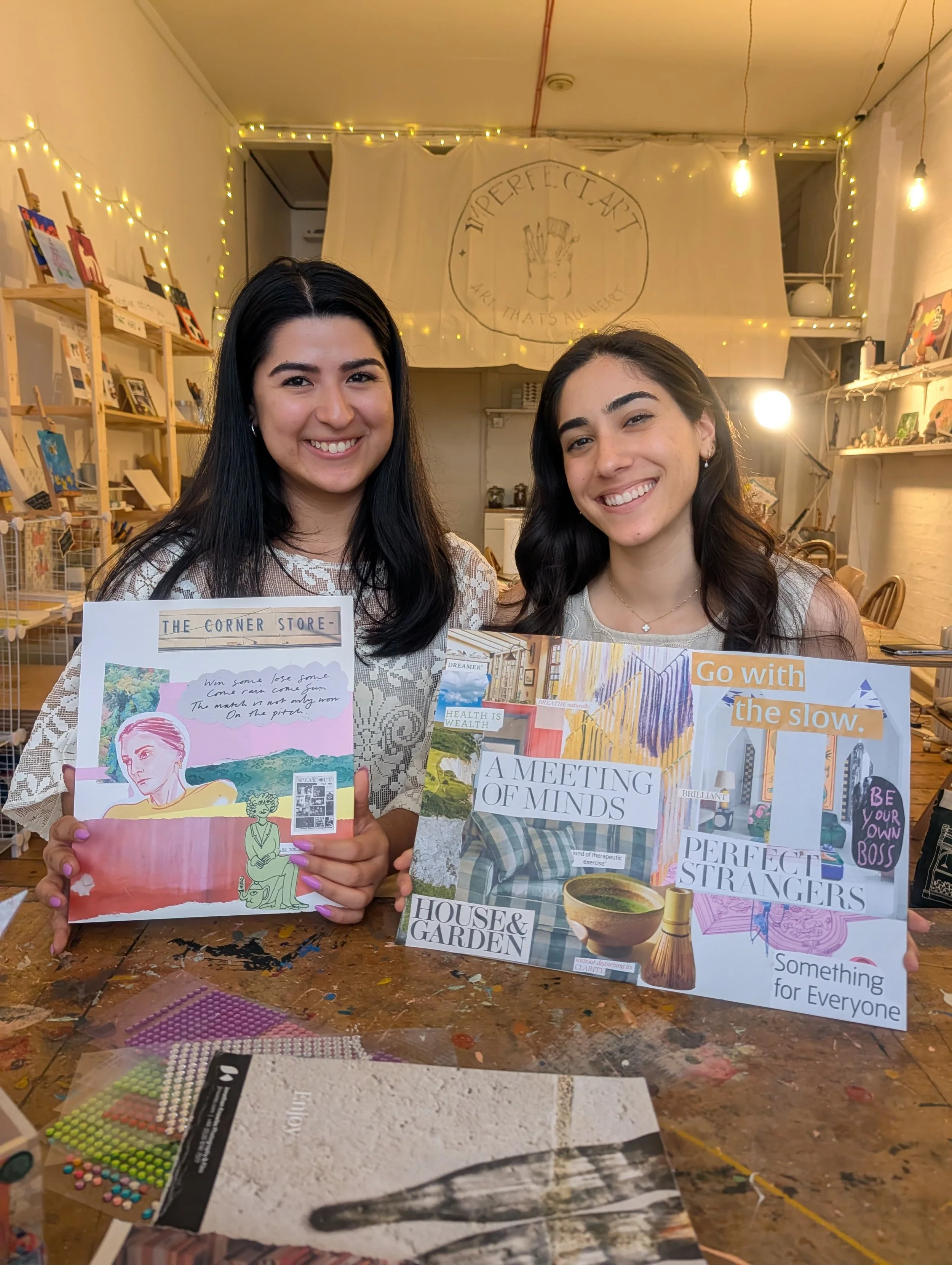 Two woman smiling and holding a board of a modern style collage, in a room warmly lit room with fairy lights. 