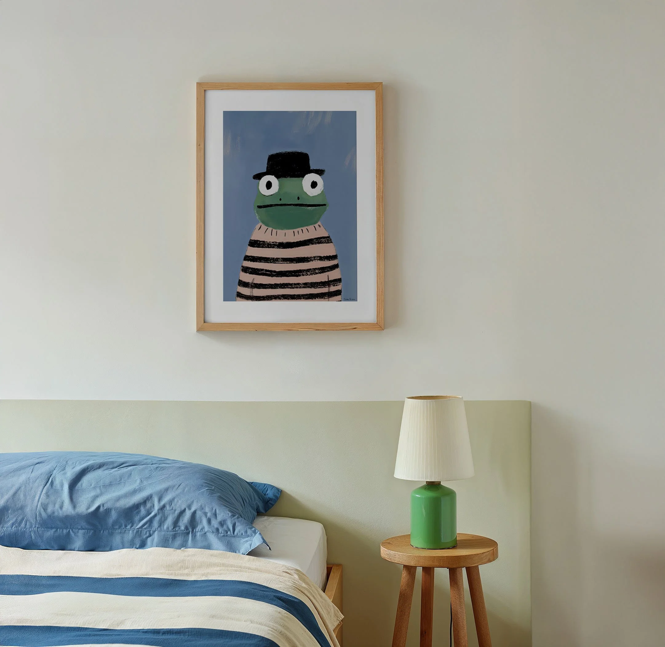 A bedroom with a framed artwork of a frog wearing a black hat and black and pink striped shirt hanging above a bed with blue bedding. There is a wooden side table with a green lamp and a white lampshade.