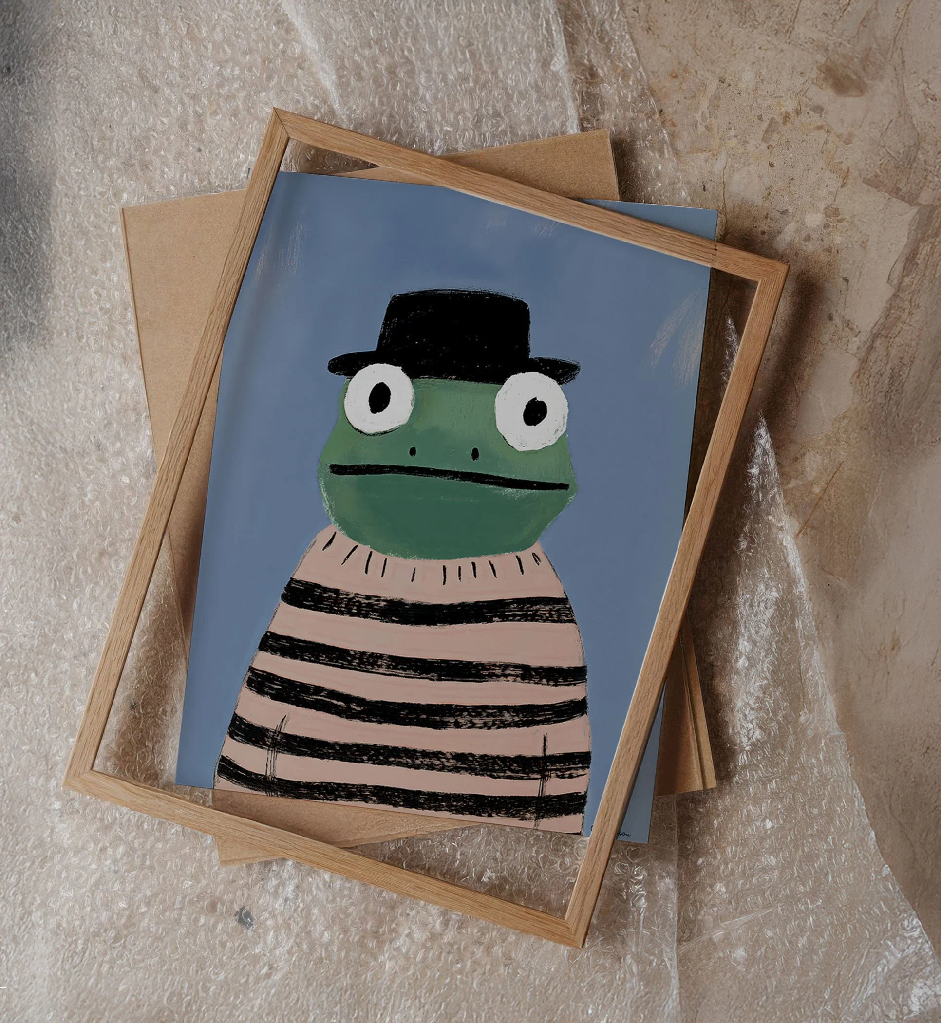A framed drawing of a green frog with large eyes, wearing a black hat and a striped sweater, on a blue background.