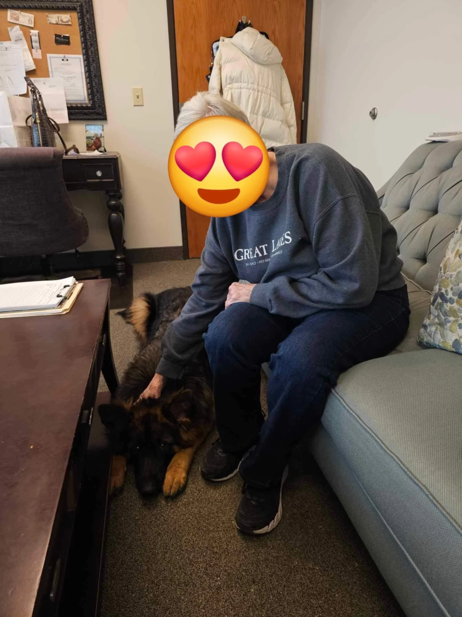 A person with a face emoji covering their face, sitting on a sofa and petting a large German Shepherd dog lying on the floor, in an office or waiting room setting.