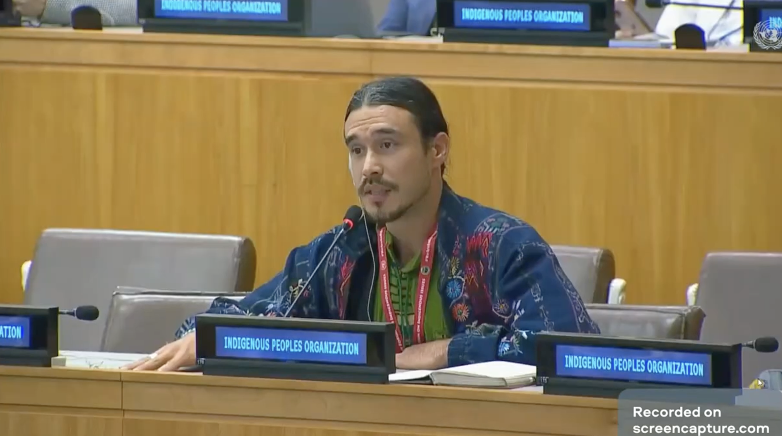 Tl'etinqox councillor Trevor Mack smiling outdoors near a river, holding a camera on a tripod over his shoulder, with a background of lush green trees and cloudy sky.