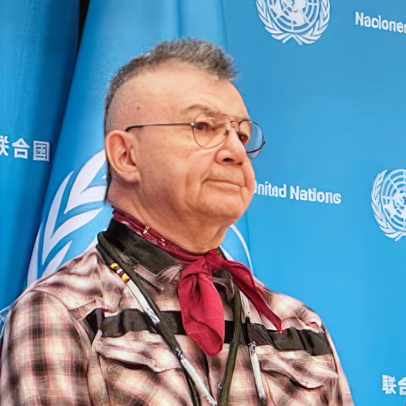 Tl'esqox chief Francis Laceese wearing glasses, a black jacket, and a red bandana, standing outdoors in front of green foliage.