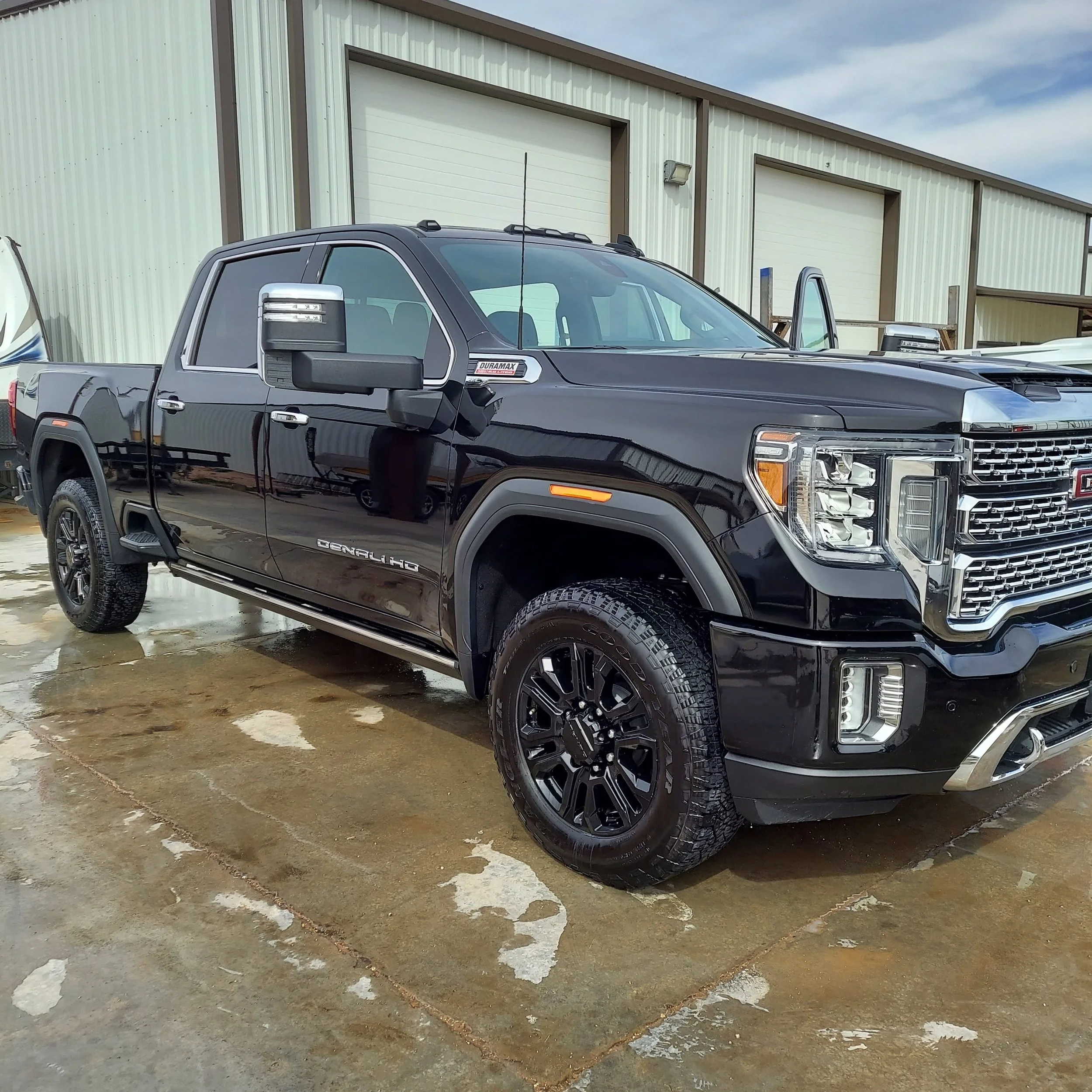 Black GMC Sierra Denali HD pickup truck parked on wet concrete in front of a beige industrial building with a closed garage door.