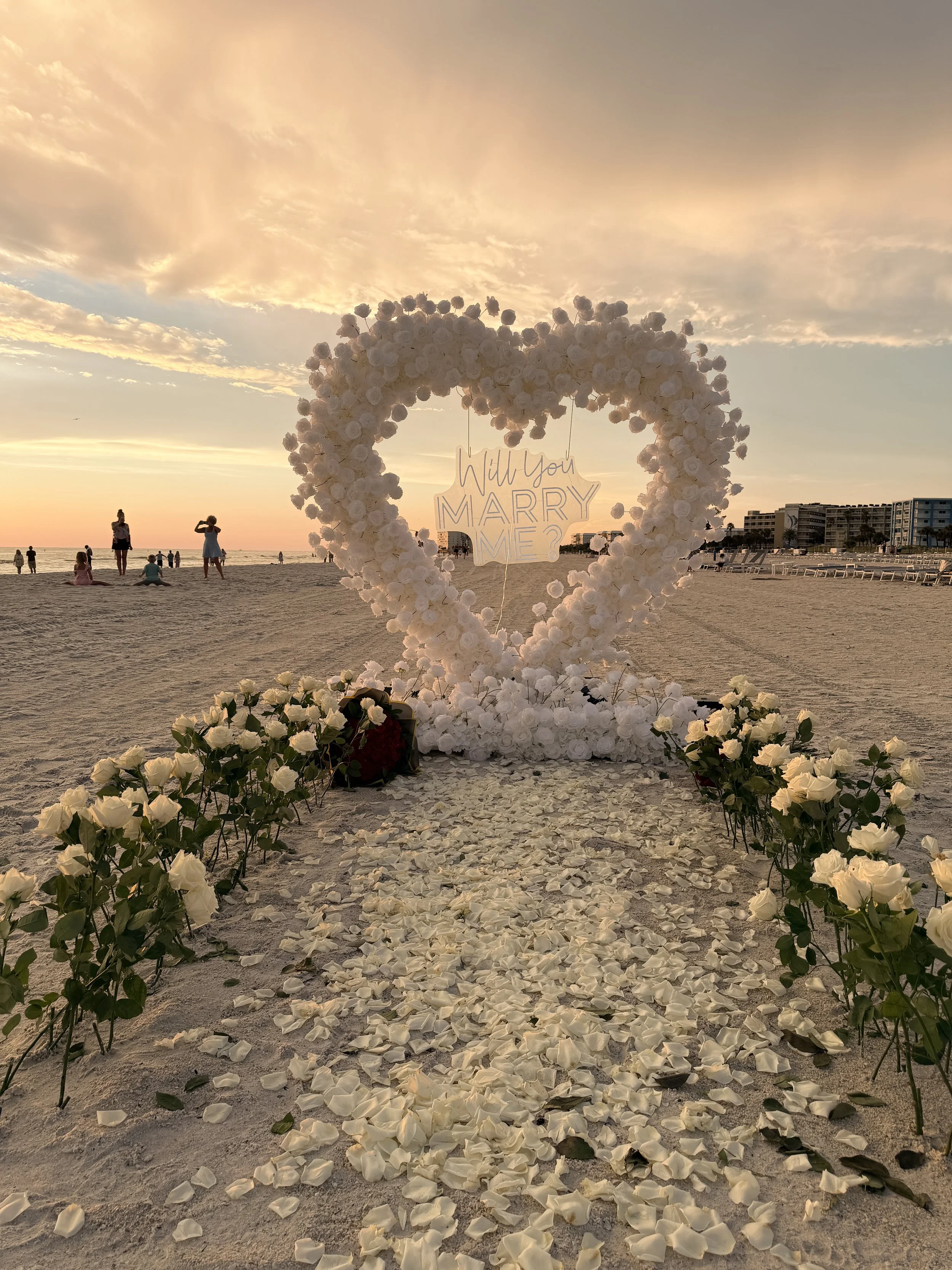 A wedding arch decorated with white roses on a beach during sunset, with a sign that says 'Will You Marry Me?' hanging inside the heart-shaped frame. White rose petals scatter on the sand leading up to the arch, withpeople and buildings visible in the background.