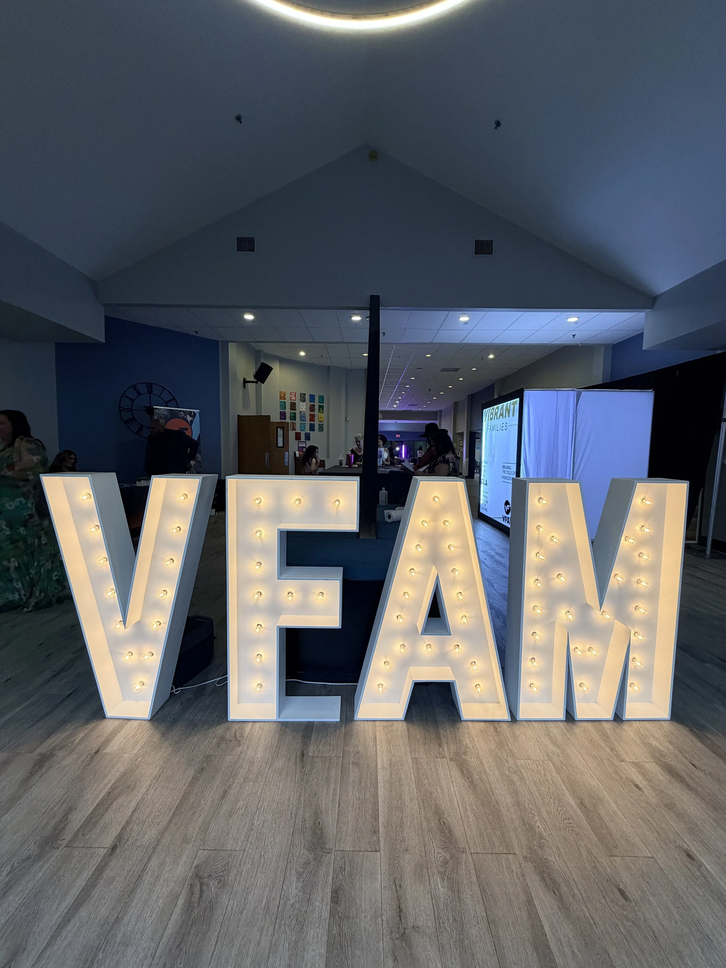 Large decorative letters (marquee letters) spelling 'VFAM' with illuminated bulbs in an indoor event space, with people and displays in the background.