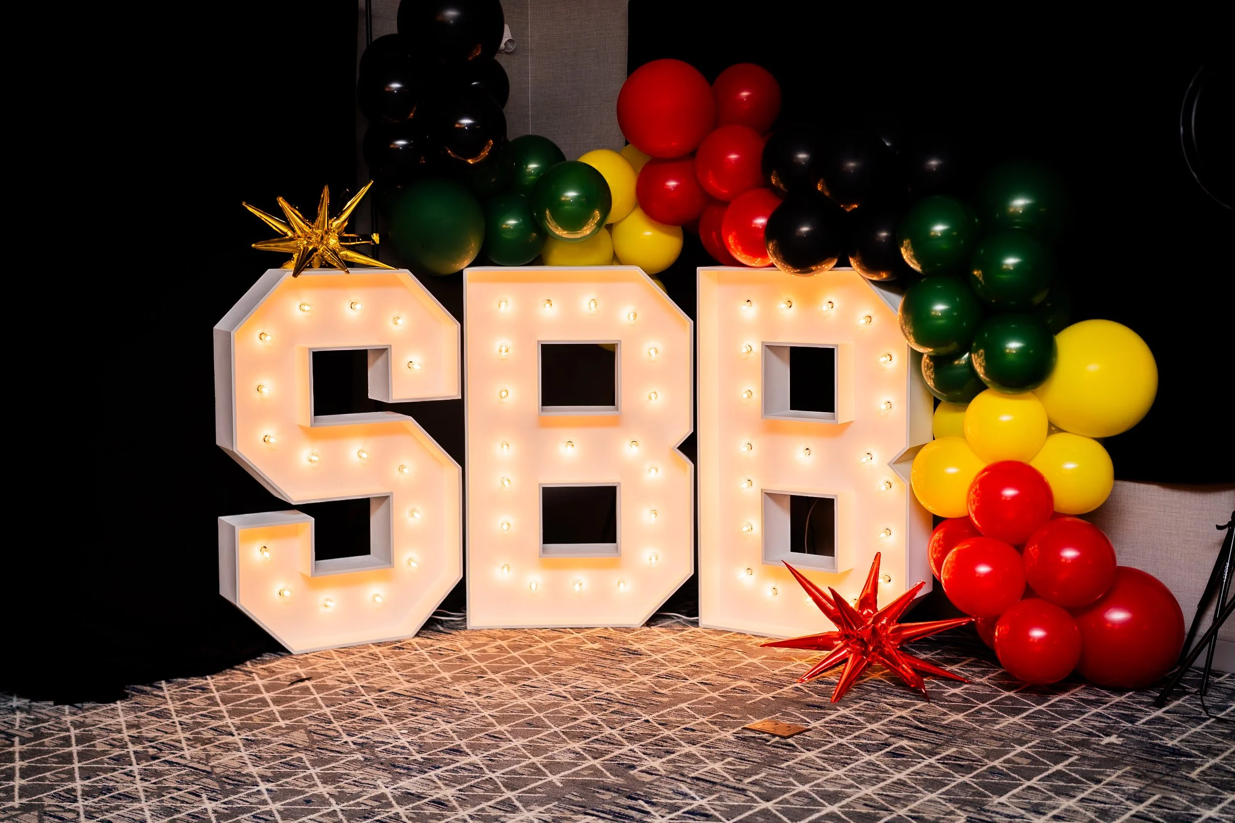 Decorative display with large illuminated, marquee letters and colorful balloons in red, yellow, green, and black, with gold and red starburst decorations on a patterned floor.