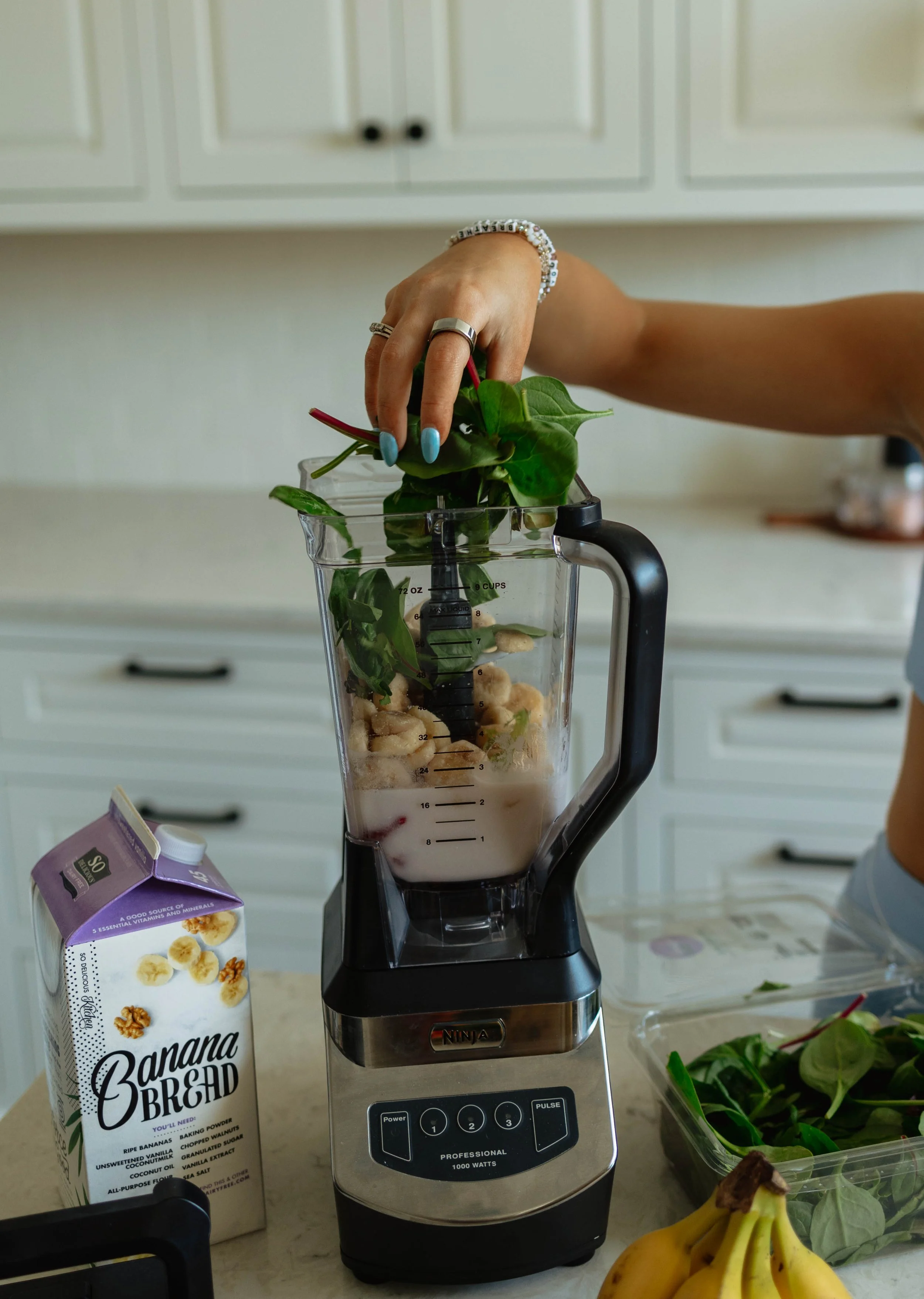 A person adding leafy greens into a blender with other ingredients, including bananas and liquid, in a kitchen. A carton of banana bread is on the counter.