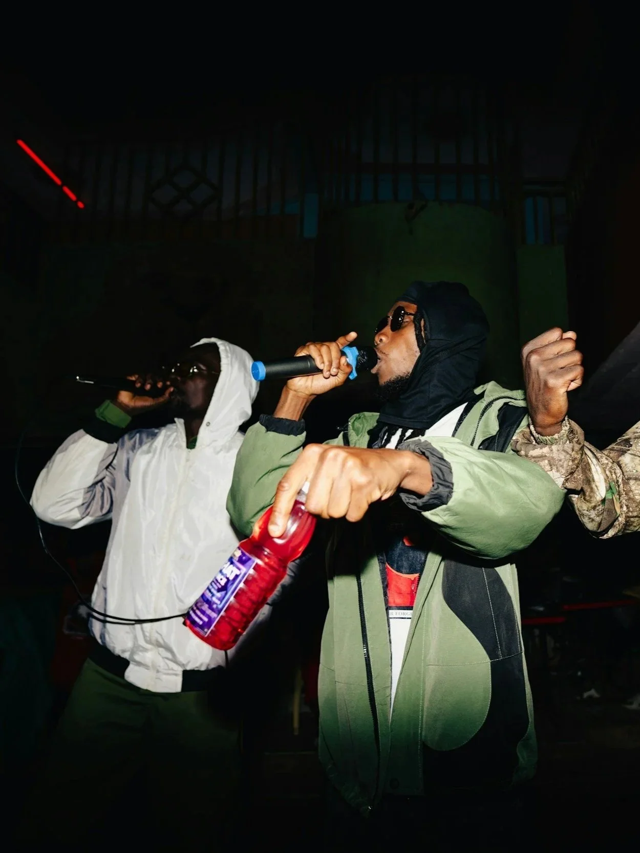 Two men wearing hoodies and sunglasses singing into microphones, one holding a fire extinguisher, in a dark indoor setting.