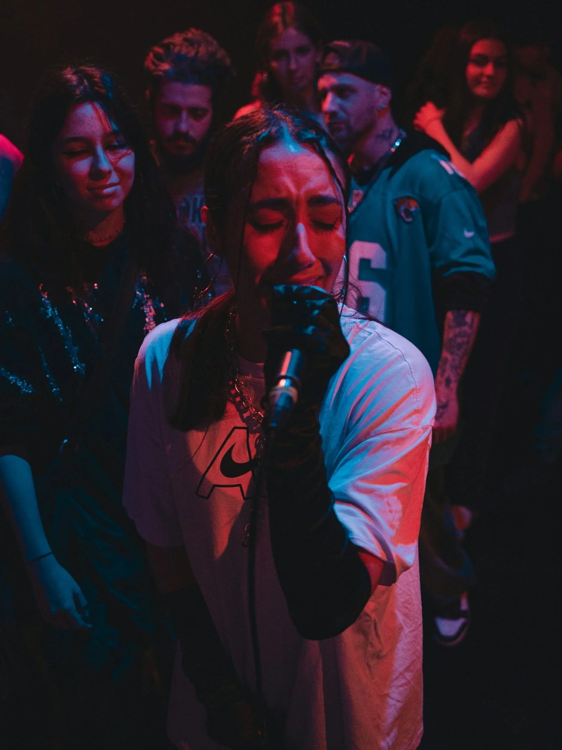 A female singer passionately performing with a microphone, surrounded by a diverse group of people in a dark, colorful setting.
