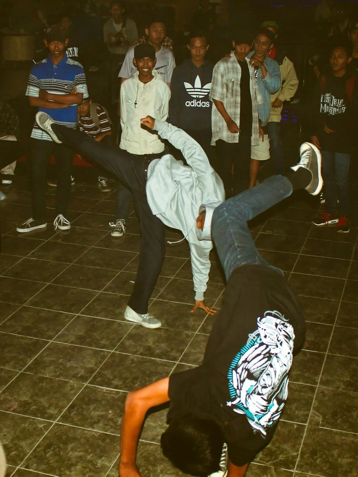 Two young men are breakdancing in the center of a group of onlookers in a dimly lit indoor space. One is performing a handstand, while the other is supporting him with their leg. The spectators are standing around watching, some with smiles and interest.