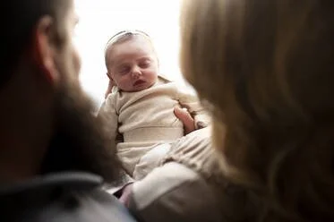 A newborn baby held by a parent, with another adult in the background.