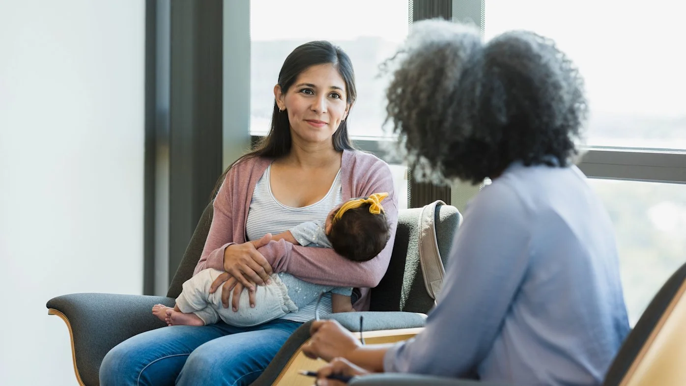 A woman holding a baby on her lap during a consultation with a therapist in a bright office.