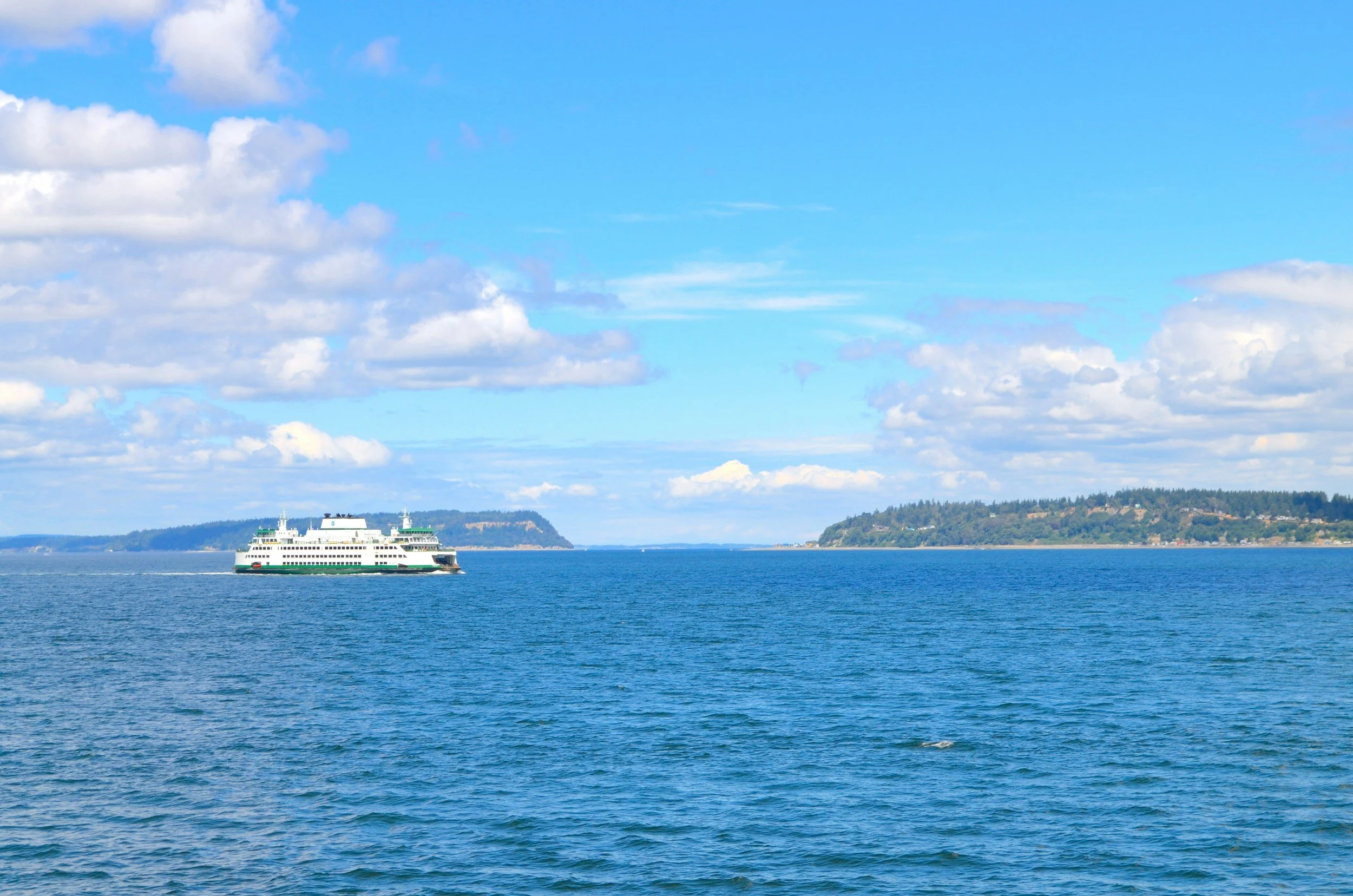 A large ferry boat sailing across a body of water with landmasses visible in the distance under a blue sky with scattered clouds.