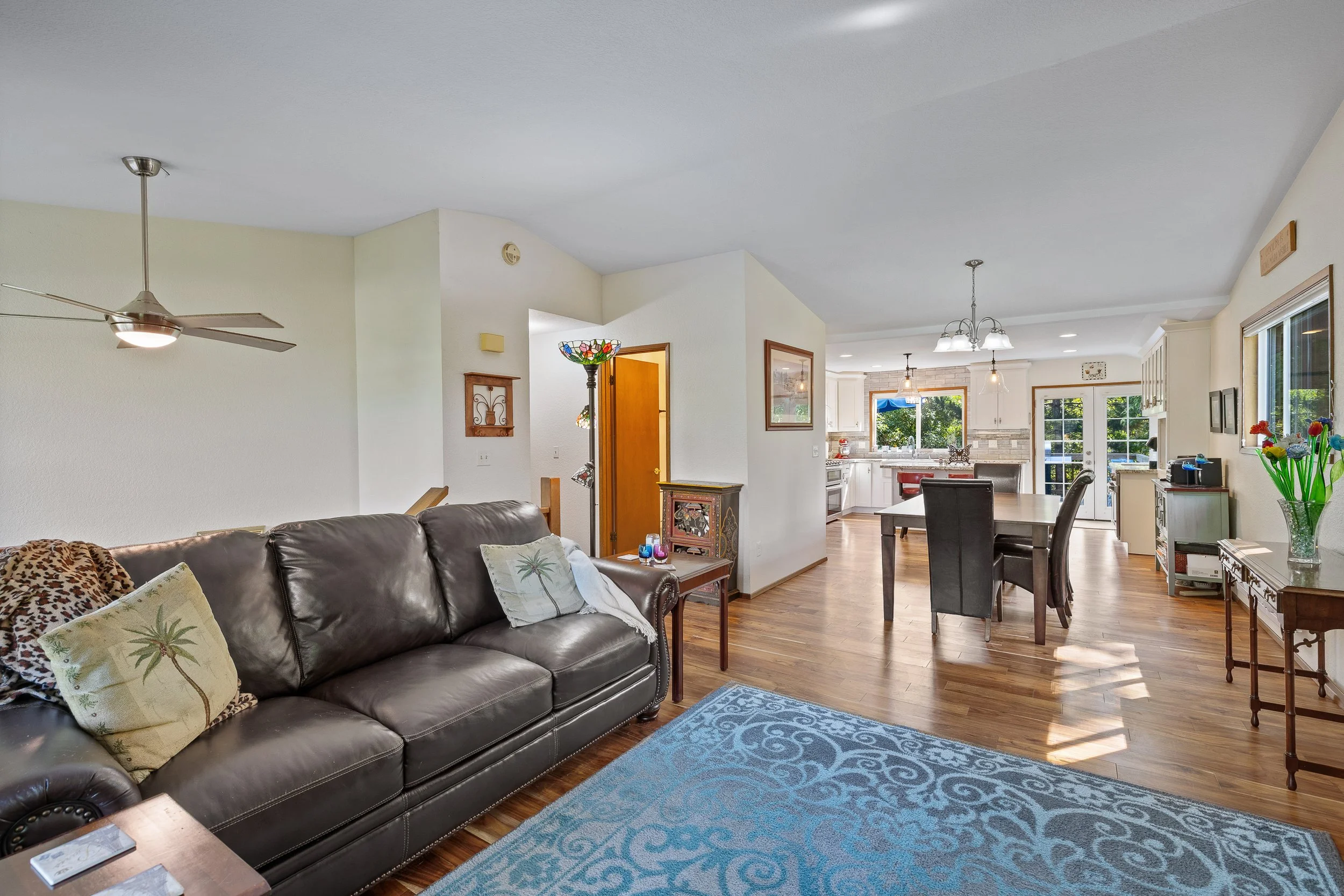 Open-concept living and dining area with hardwood floors, leather sofa, area rug, wooden table, kitchen with white cabinets, stainless steel appliances, and large window showing greenery outside.