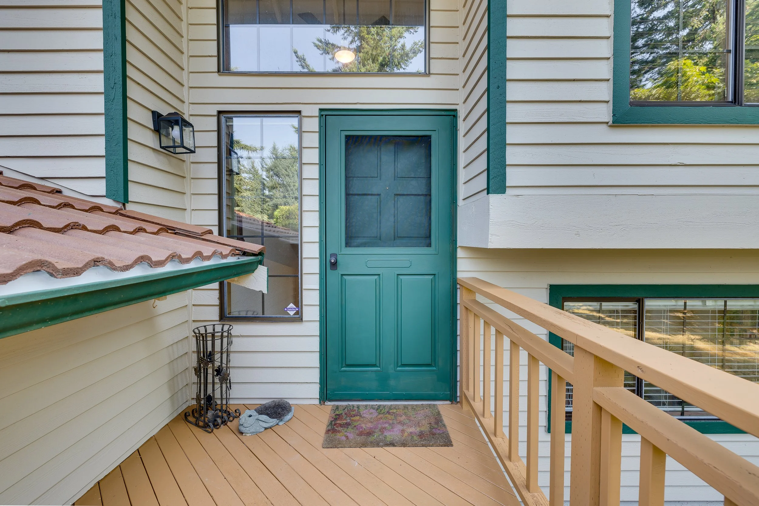 Front porch of a house featuring a teal door, large windows, a small doormat, and outdoor lighting, with a wooden railing and siding.