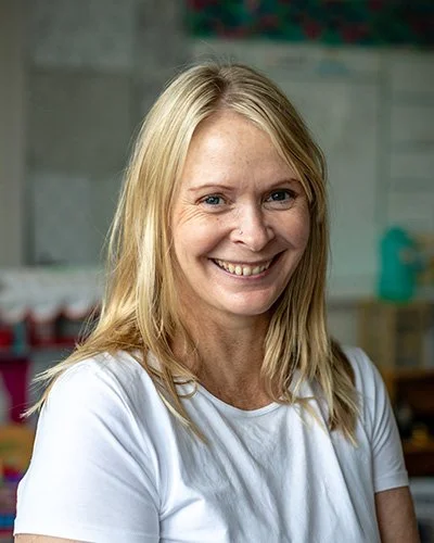 A smiling woman with blonde hair and wearing a white shirt in a classroom setting.