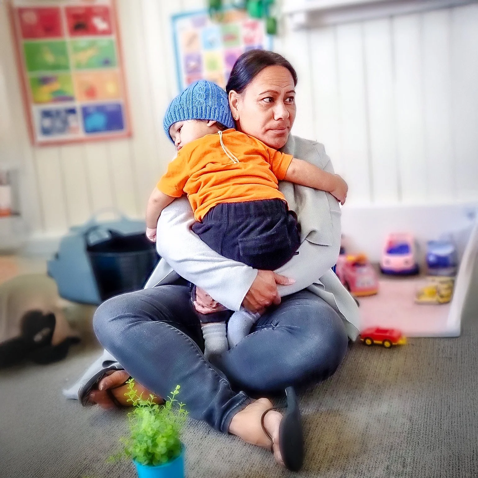 A woman sitting on the floor holding a young boy in her arms inside a room with colorful toys and posters, a small potted plant in the foreground.