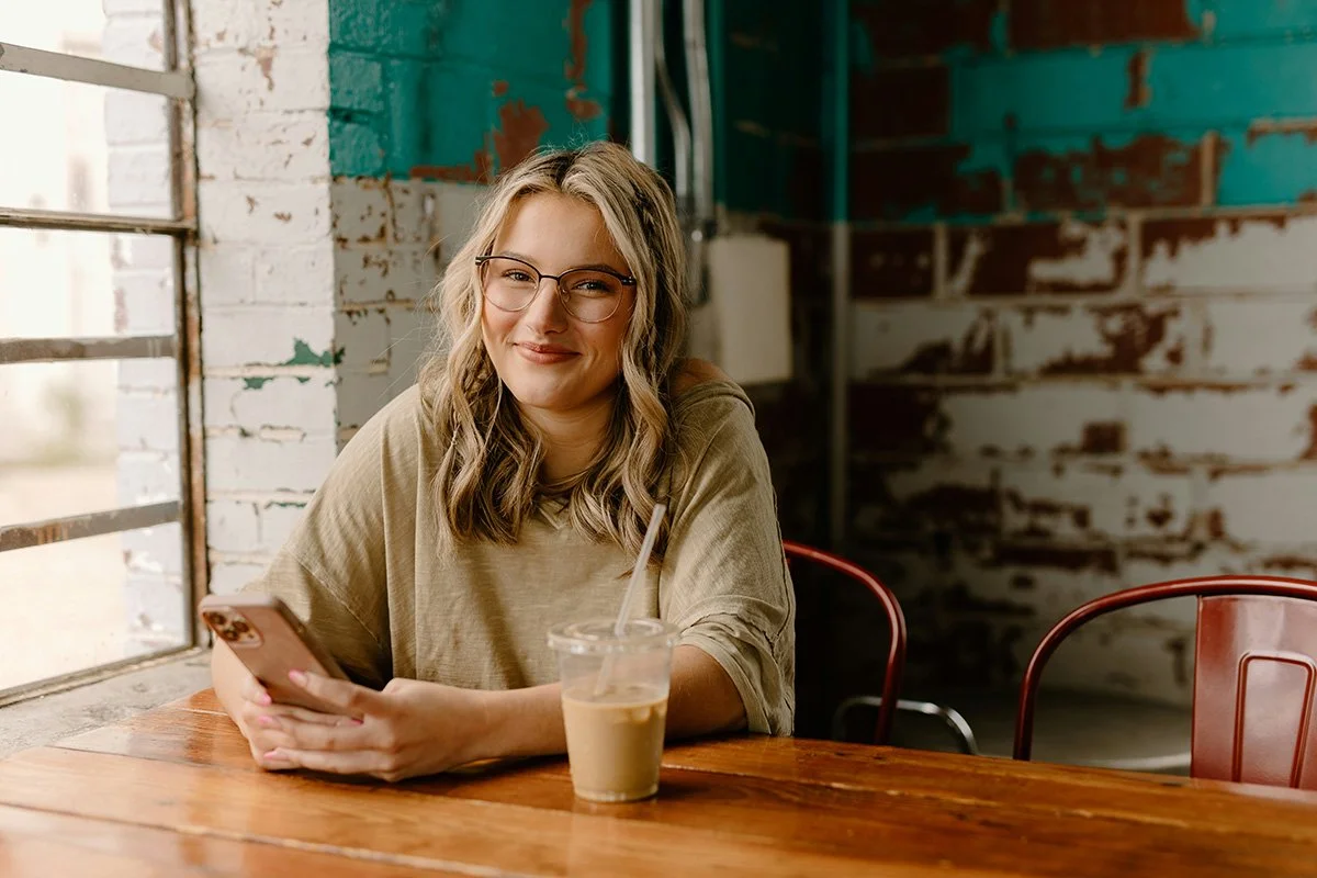 A woman with blond, wavy hair and glasses sitting at a wooden table, holding a smartphone, with a plastic cup of iced coffee in front of her, smiling at the camera in a rustic cafe with white and teal walls.