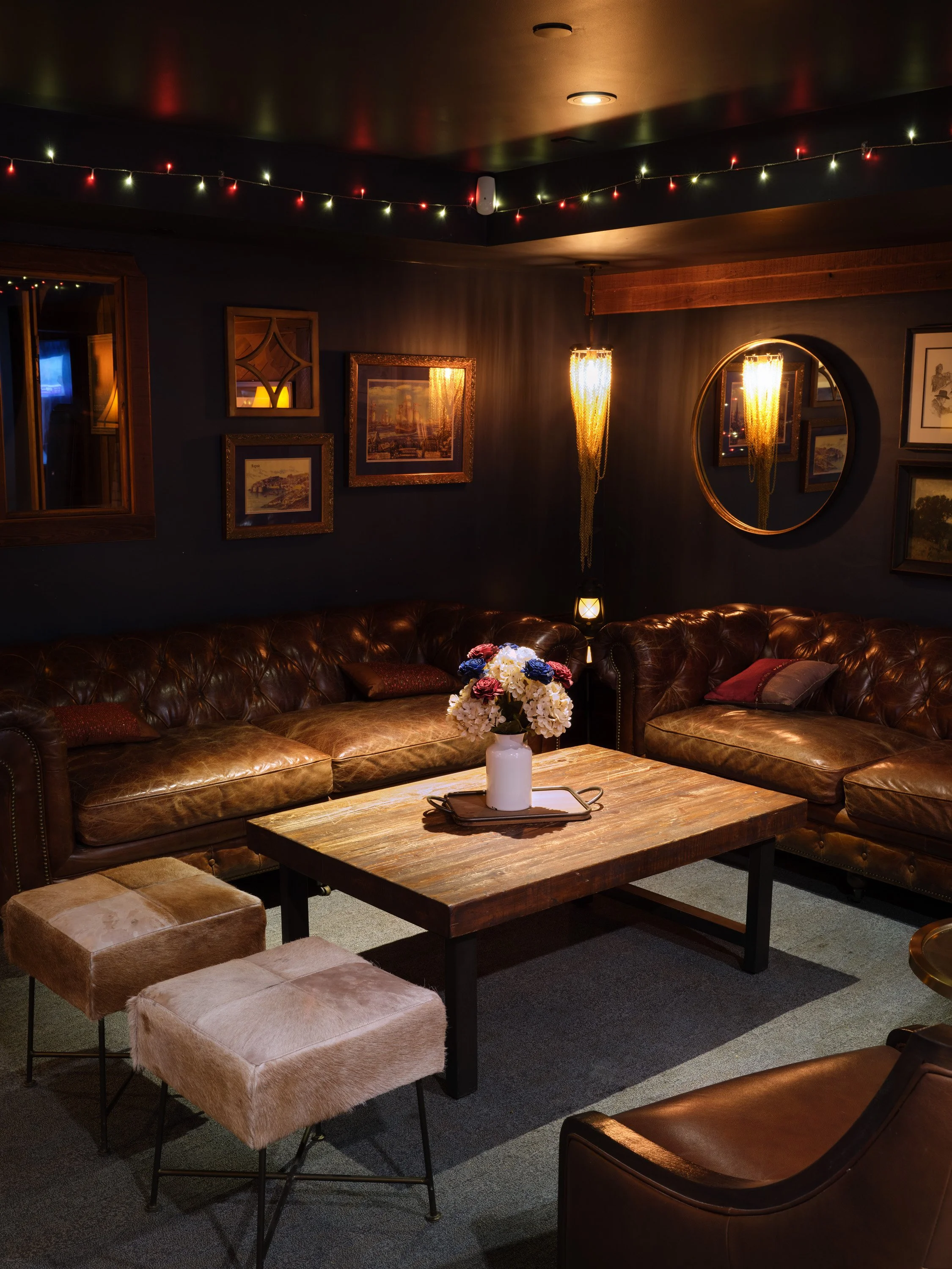 Dimly lit lounge area with two leather sofas, a wooden coffee table with a white vase of flowers, framed artwork on dark walls, decorative string lights on the ceiling, a round mirror, and a hanging light fixture.