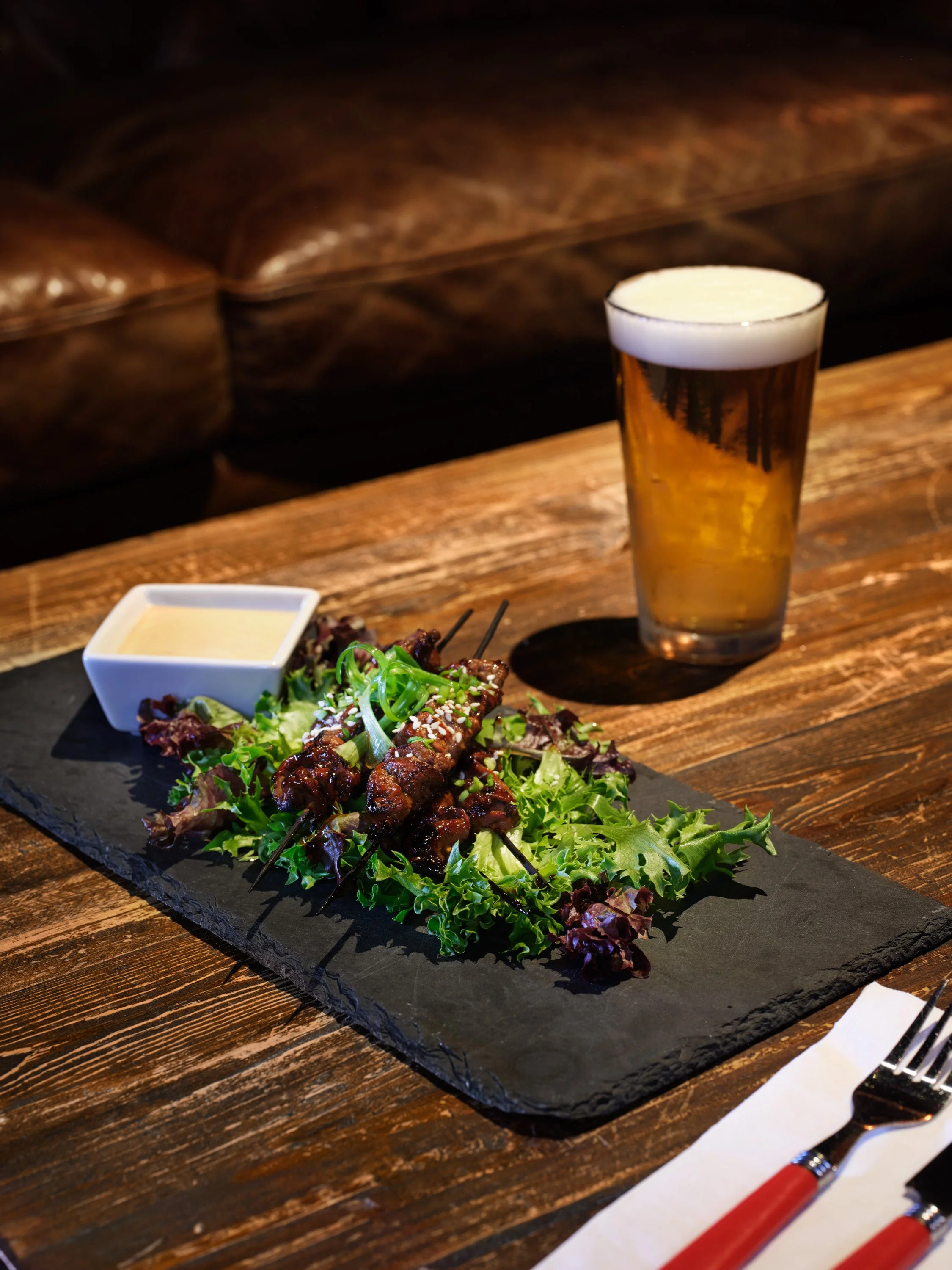 A glass of beer with foam on top, a plate of grilled meat on skewers with green leafy lettuce and a small bowl of dipping sauce, set on a black slate serving board on a wooden table, with a white napkin and red-handled utensils nearby.