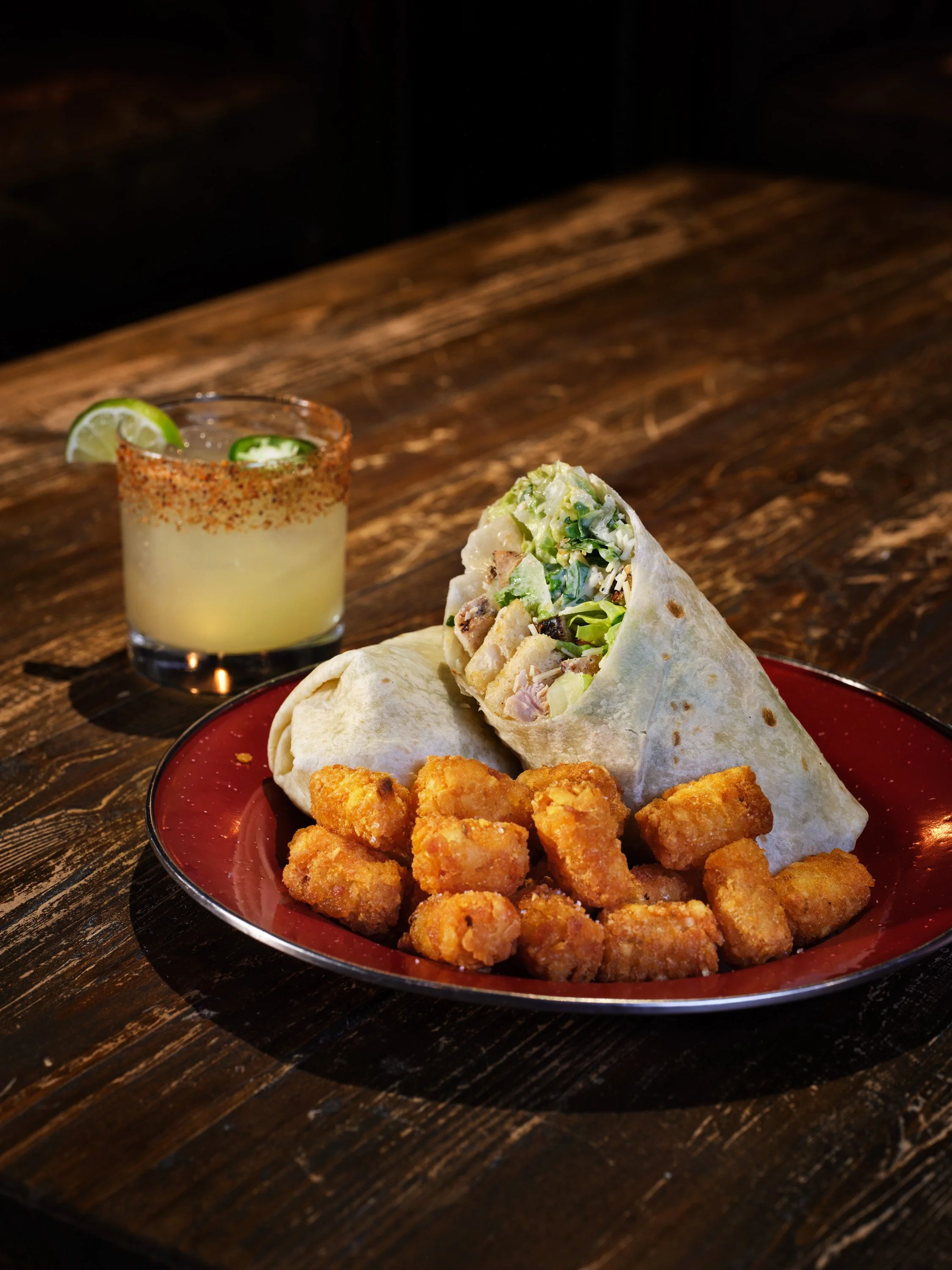 A plate with chicken nuggets, a chicken caesar wrap, and a side of fried tater tots, with a drink garnished with lime and jalapeño slices.